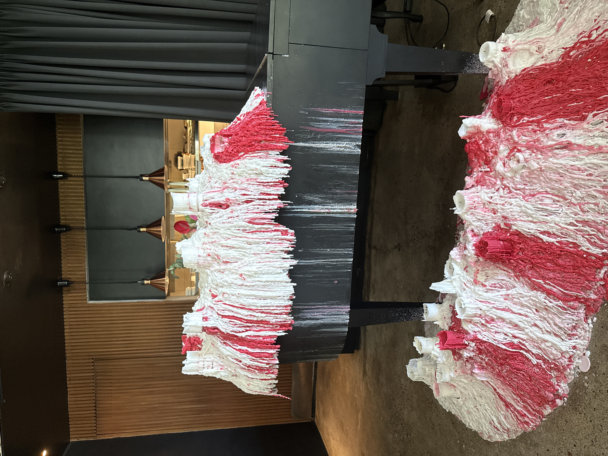 The image shows an art installation in Kuta Selatan, Indonesia. A black grand piano is covered with melted white and red candles. The melted wax drips down the sides of the piano and onto the floor. A large puddle of melted wax, also with white and red candles, is spread out on the floor in front of the piano. The installation is in a room with a dark ceiling, wood-paneled walls, and a gray curtain on the right side. There are three hanging lamps with copper-colored shades above the piano.