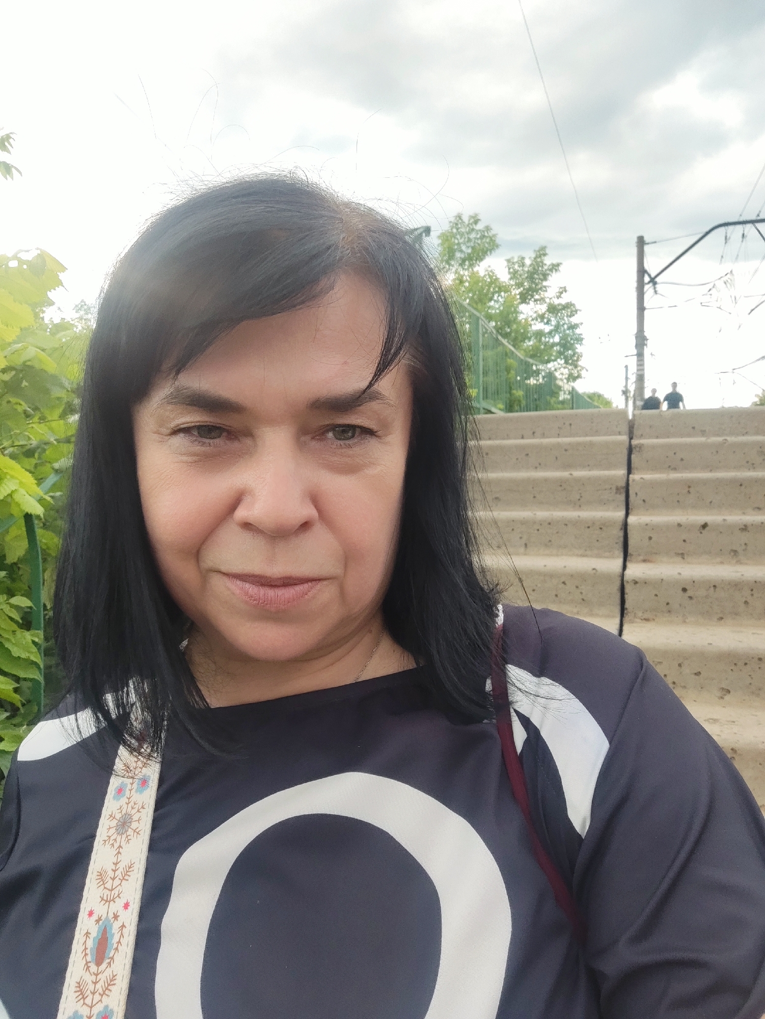 A woman with shoulder-length black hair and brown eyes is looking at the camera in an outdoor setting. She is wearing a dark-colored shirt with a large white graphic design. A patterned strap of a bag is visible across her chest. In the background, there are concrete stairs leading upwards, a green fence, trees, and utility poles with wires. Two people are visible in the distance on the stairs. The sky is cloudy.