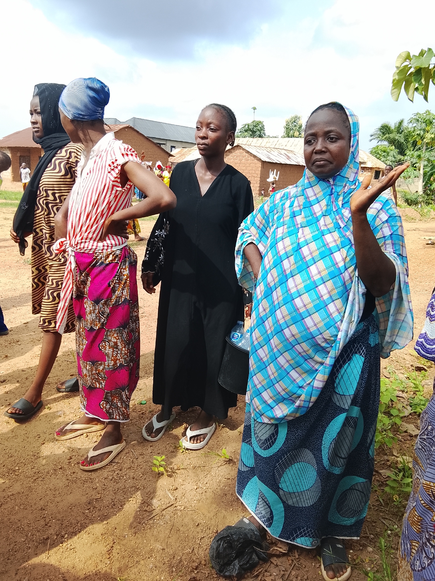 The image depicts a group of women in an outdoor setting, likely in Akwanga, Nigeria. The foreground features four women, three of whom are clearly visible from the waist up. They are standing on a dirt ground. The woman on the far right wears a blue and white checkered headscarf and a blue and white checkered garment. Her right arm is raised, and she appears to be speaking or gesturing. To her left, a woman in a black headscarf and black dress looks to her left. Next to her is a woman with a blue and white striped headscarf, wearing a red and white striped top and a pink patterned skirt. The woman on the far left is partially obscured by the woman in the striped top. In the background, several rustic, red-brick buildings with corrugated metal roofs are visible. There are also trees and foliage, suggesting a rural or semi-rural environment. The sky is partly cloudy, indicating it might be daytime. The overall scene appears to be a casual gathering or a public interaction.