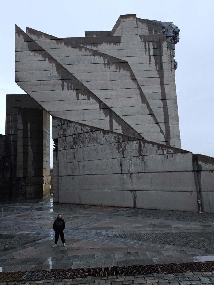 The image depicts the "Founders of the Bulgarian State Monument" in Shumen, Bulgaria, under an overcast sky. The monument is constructed from large, angular concrete blocks arranged in a tiered, brutalist architectural style. Dark vertical streaks, indicative of weathering and water runoff, are visible on the grey concrete surfaces. A large, sculpted head, possibly a lion or stylized human figure, is visible on the upper right section of the main structure. To the left, another section of the monument features two tall, parallel concrete slabs forming a narrow passageway.
One adult male is standing in the lower-center of the frame, facing forward. He is wearing a dark hooded jacket, dark trousers, and white athletic shoes. The ground is paved with irregular grey flagstones and cobblestones, which appear wet and reflective, suggesting recent precipitation. A metal drainage grate is present in the immediate foreground. No active interactions are occurring; the individual is stationary.