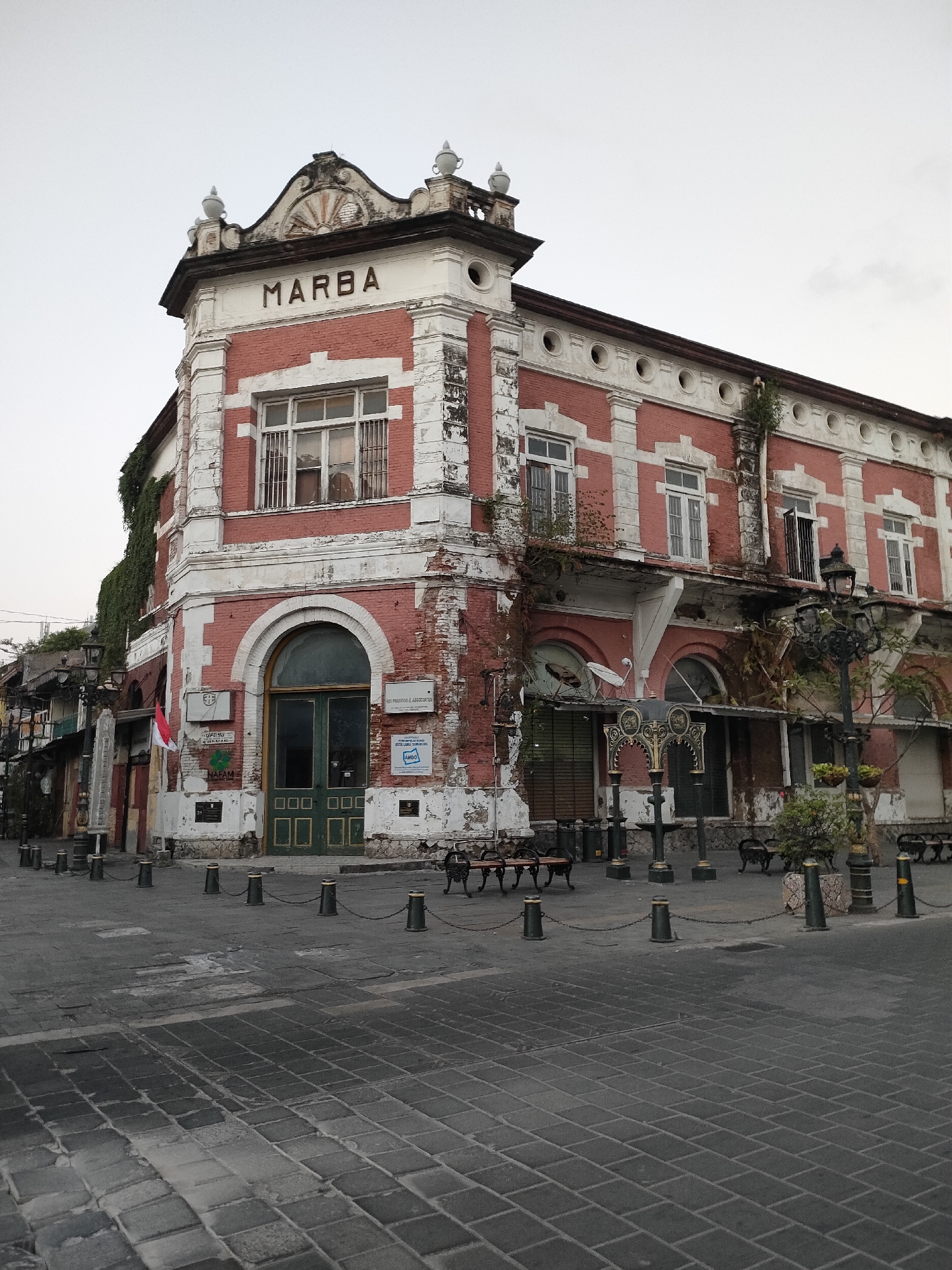 This is a medium-angle, eye-level outdoor shot of the Marba Building in Semarang, Indonesia. The building, with its red brick facade and white accents, exhibits signs of age and disrepair, with peeling paint and overgrown vines. The building has a prominent "MARBA" sign at its peak, and an arched green double door on the ground floor. The street in front of the building is paved with cobblestones, and lined with bollards and chains. Benches are placed along the street. The sky is overcast. There are no people visible in the image. The scene appears to be in a historical district of Semarang.