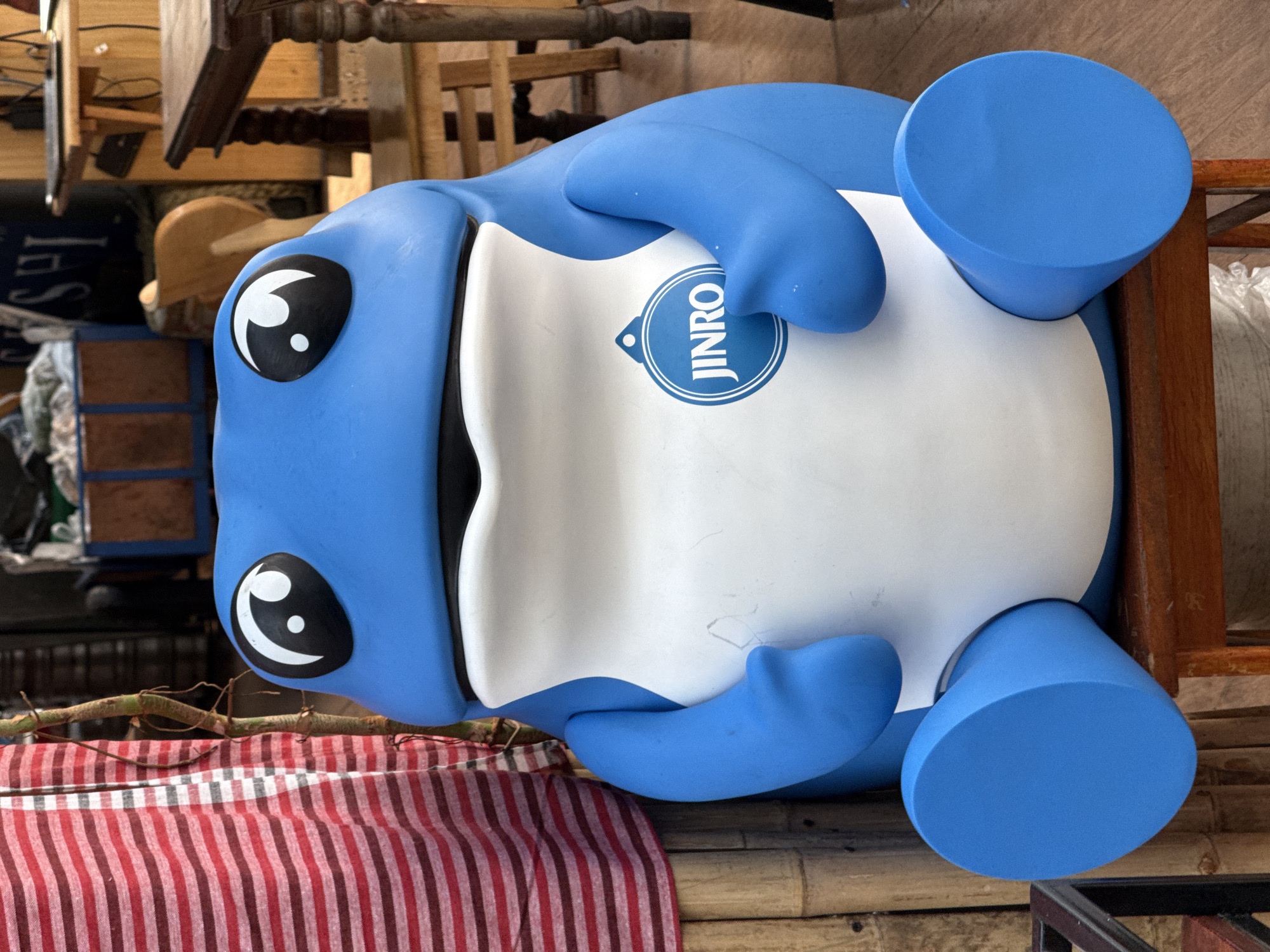 The image shows a large, inflatable, blue and white frog-like figure with the "JINRO" logo displayed on its white belly. It has large, cartoonish eyes. The figure is lying on its back on what appears to be a bamboo mat. A red and white striped cloth is partially visible beneath the figure. The background suggests an indoor or covered space with stacked furniture and various objects. The setting might be a shop or storage area.
It is impossible to definitively determine the precise location in Bangkok, Thailand from the image alone.