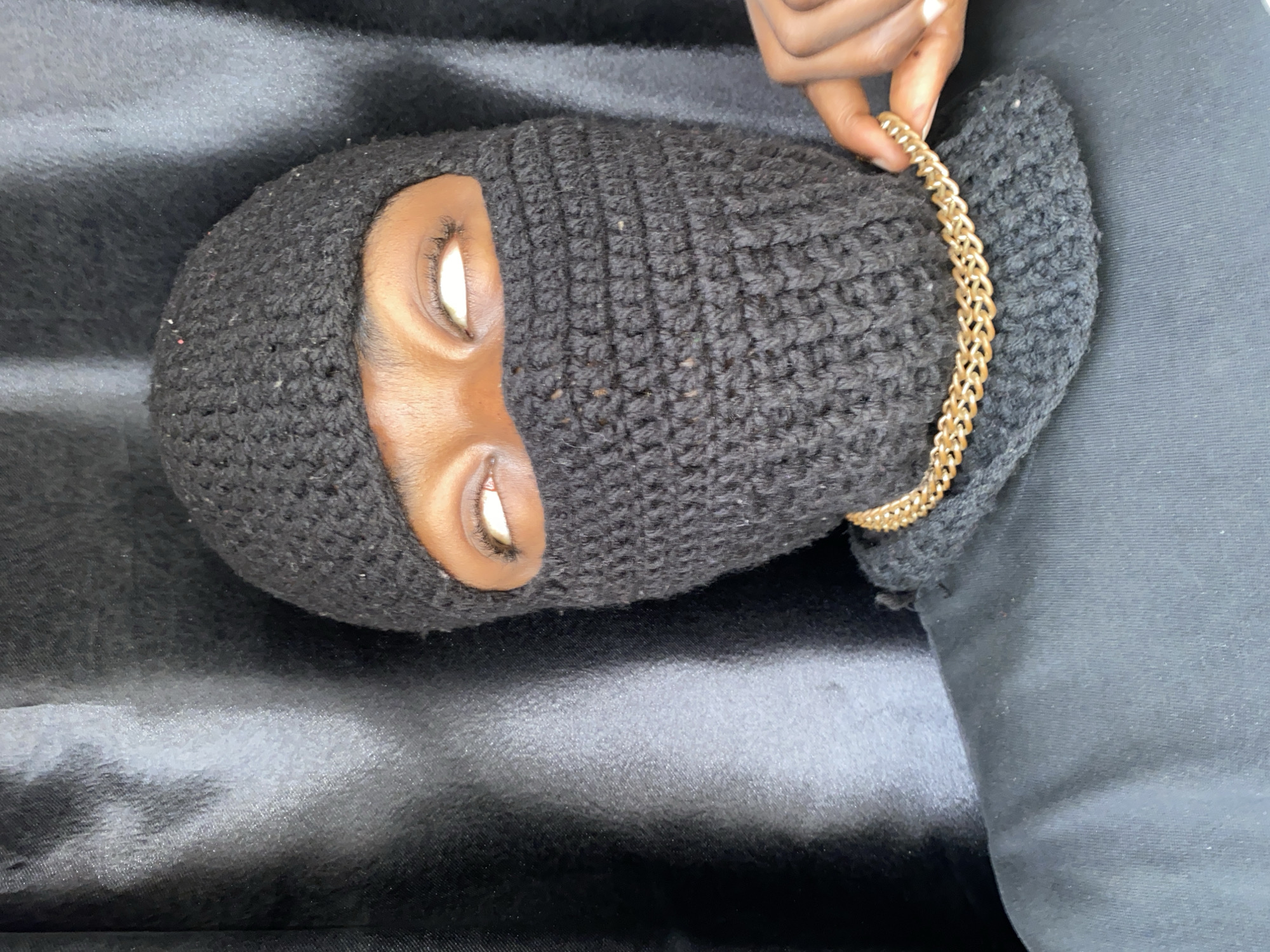 A person wearing a black balaclava lies on a dark, possibly satin or velvet, surface. Their eyes are visible through the openings in the balaclava. A hand holds a gold chain necklace near the person's head. The background is out of focus, but some dark clothing is visible. There is no indication that this photo was taken in Barakin Akawo, Nigeria, or any specific location.