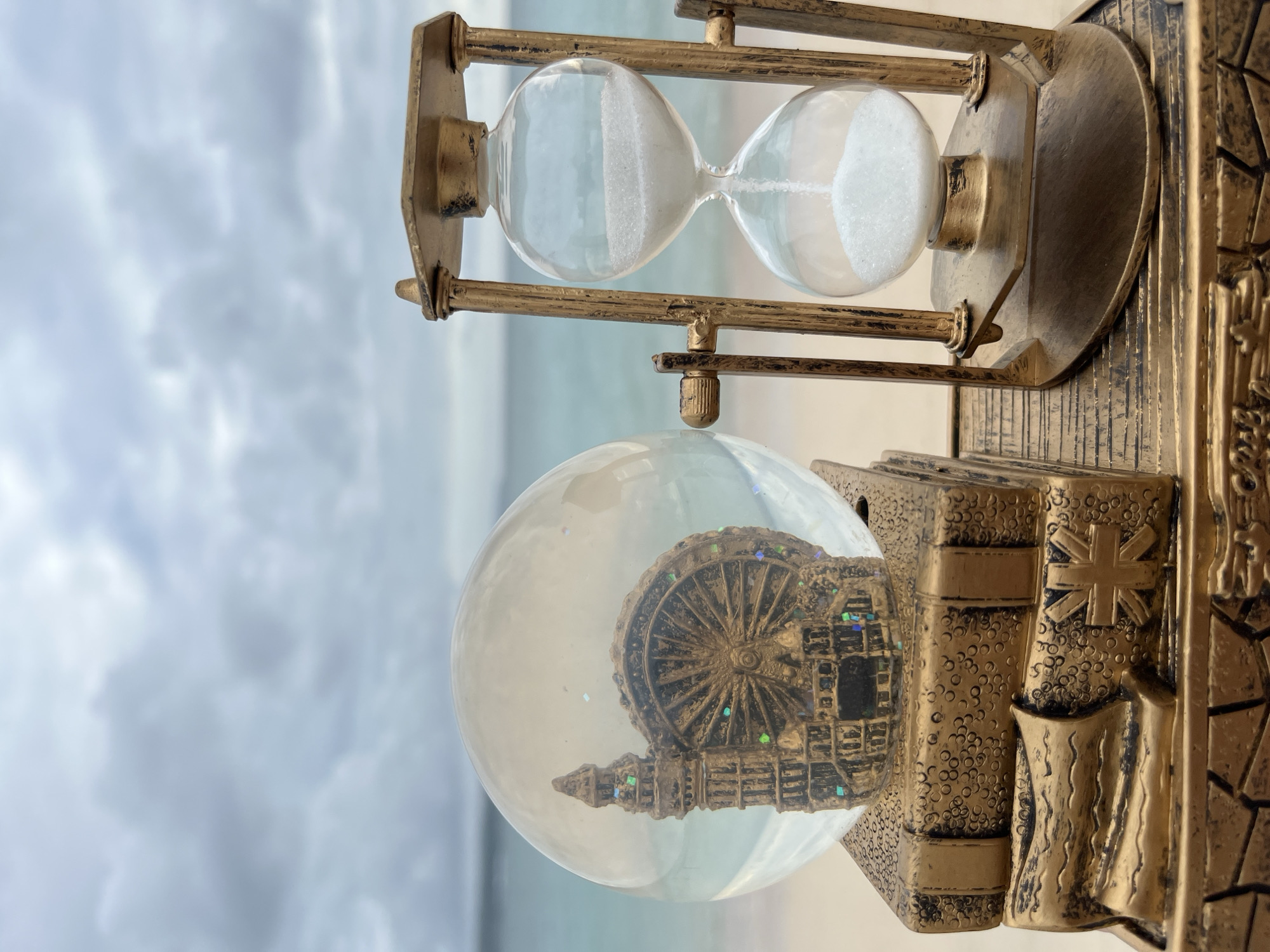 In Kuta, Indonesia, a golden hourglass stands beside a snow globe containing a miniature Ferris wheel and a tall tower, both resting on a stack of decorative gold books, against a backdrop of a cloudy sky and a serene ocean. The sand in the hourglass is evenly distributed between its two glass bulbs, and the snow globe's contents sparkle with glitter. The scene is meticulously detailed, with the gold finish of the hourglass and book decorations exhibiting a subtle textured pattern. The ocean stretches out in the distance, its waves gently lapping at the sandy shore. The overall impression is one of quiet contemplation, a juxtaposition of iconic city landmarks with the tranquil beauty of nature.
