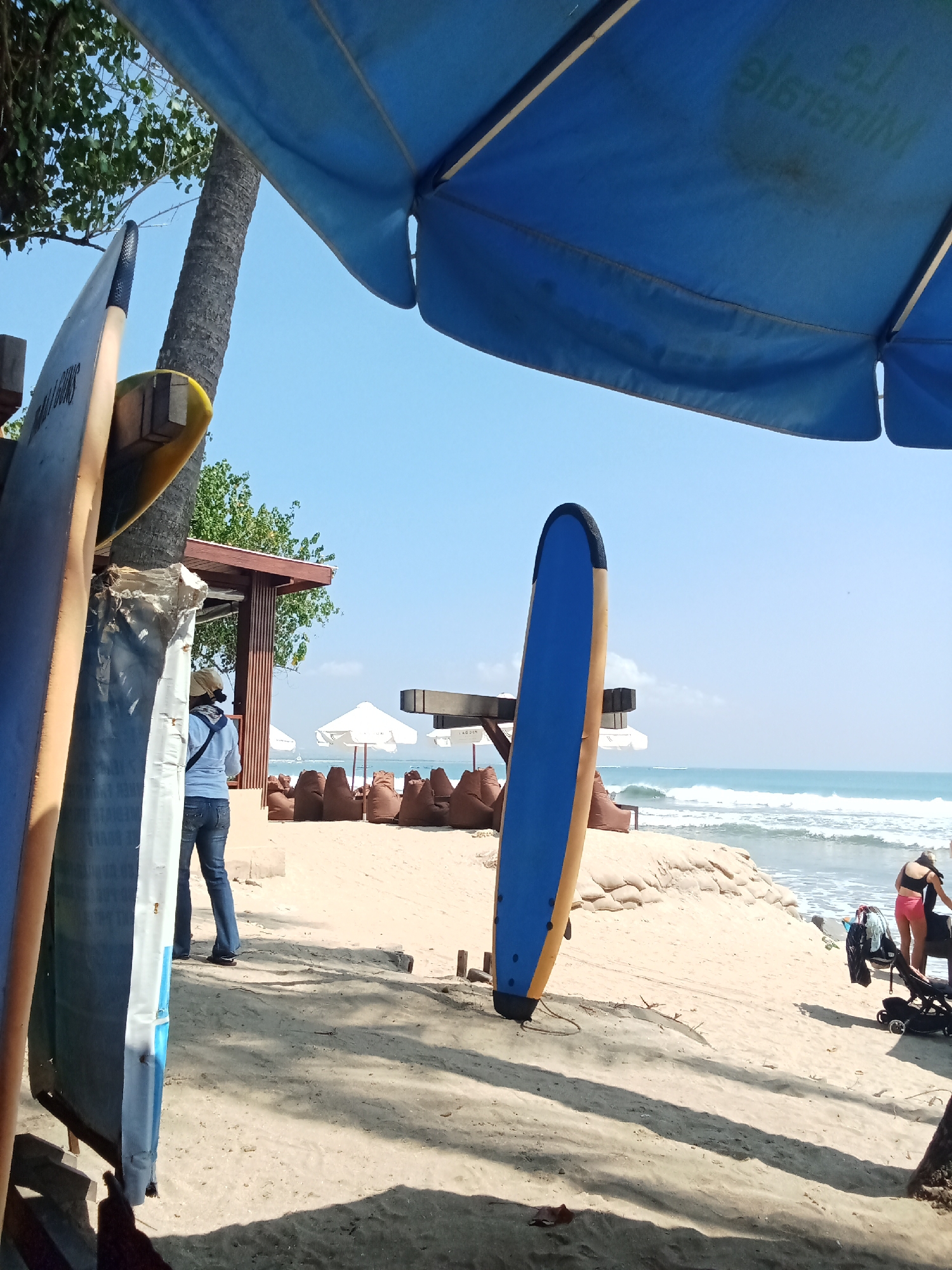 The image depicts a beach scene under a bright, clear blue sky. In the foreground, two surfboards are propped up, one with a blue and yellow design and another mostly obscured, appearing white with blue accents. To the left, a person wearing a light-colored hat, a blue shirt, and jeans stands facing away from the camera, with a dark bag slung over their shoulder. Behind them, a palm tree with a rough trunk rises towards the sky.
Further into the scene, a beach area is set up with numerous beanbag chairs in a warm, brown color, clustered in front of a low structure with a wooden roof. White umbrellas are visible in the background, providing shade. The ocean stretches out to the horizon, with moderate waves breaking on the shore. In the middle ground on the right, a person in a pink top and shorts is attending to a stroller, with laundry or bags placed on it. The sand is light brown and textured, with some areas of shadow cast by unseen objects. The overall impression is a relaxed, sunny day at the beach, possibly a resort or a popular surf spot.