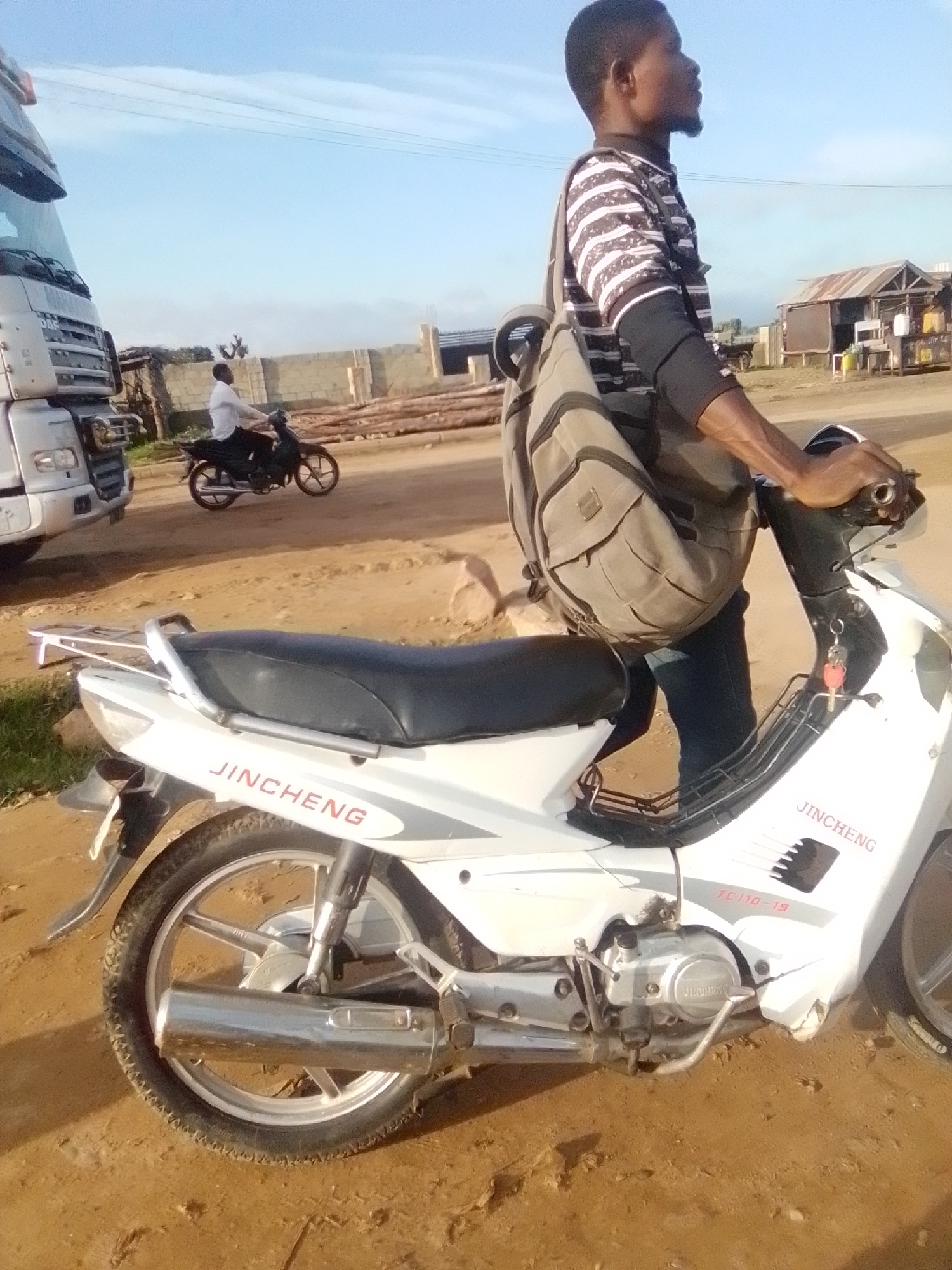 The image captures a side profile of a man on a white JinCheng motorcycle. He is wearing a horizontally striped t-shirt and dark blue jeans, with a beige backpack slung over his left shoulder. The motorcycle has a black seat and a chrome exhaust pipe. In the background, a large white truck is visible on the left, and a person on another motorcycle is seen in the distance. The environment appears to be a dusty, unpaved area, possibly a roadside or construction site, with a clear sky overhead. This scene is located in the city of Kwadon, Nigeria.