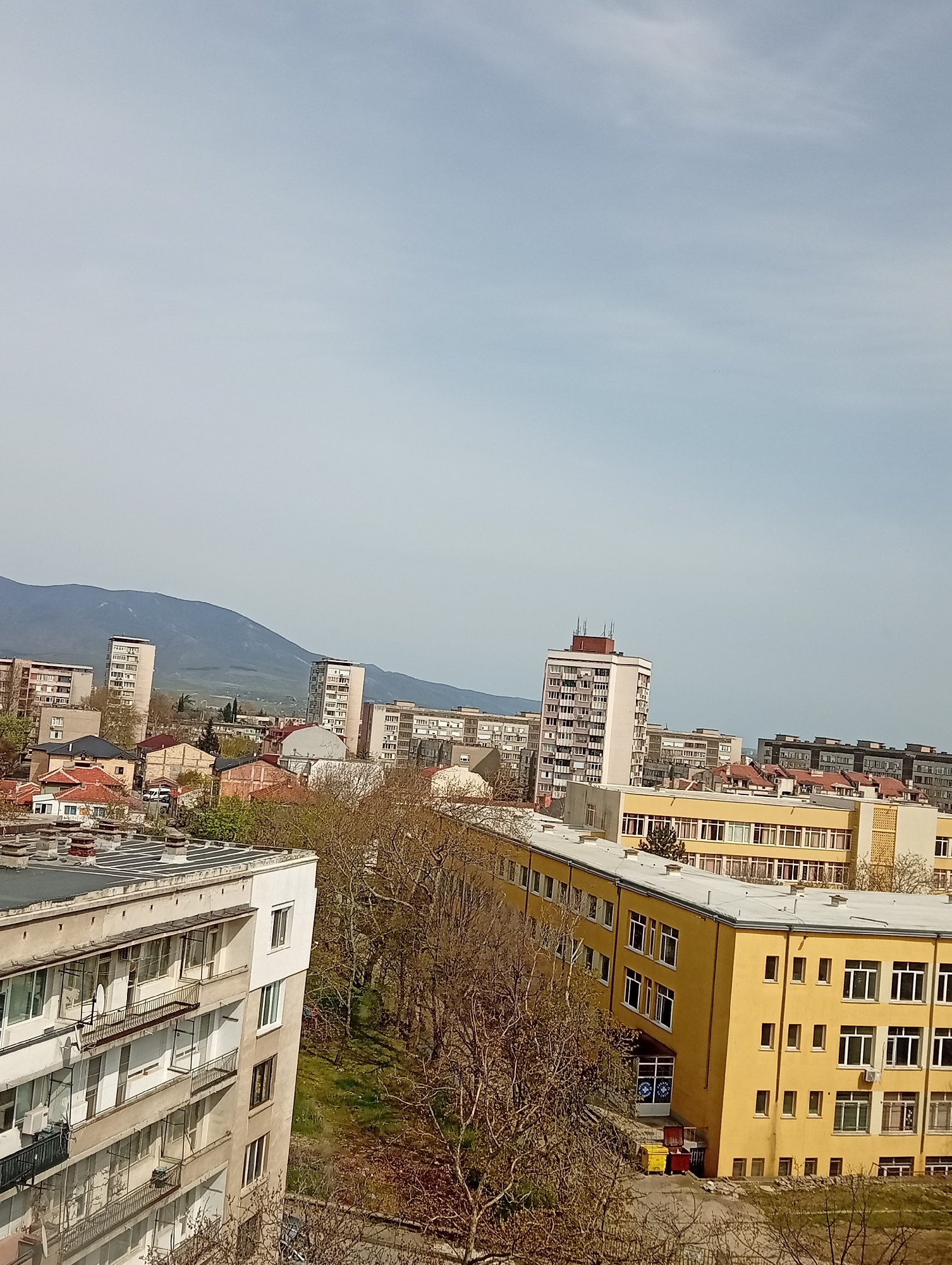 The image shows an urban landscape in Unknown, Unknown, featuring several buildings, a mountain range in the background, and a blue sky. The scene is viewed from an elevated position, possibly from a high-rise building.
Several buildings are visible, including apartment blocks and a large yellow structure with many windows, resembling a school or public building. A cluster of bare trees is situated between the buildings, suggesting it might be late fall or winter. The mountain range in the distance is a light bluish-gray color and appears to be partly obscured by a light haze.
The sky is light blue with some areas of cloud cover. No people or animals are clearly visible in the scene. The photo is taken during the daytime, with soft lighting casting long shadows.