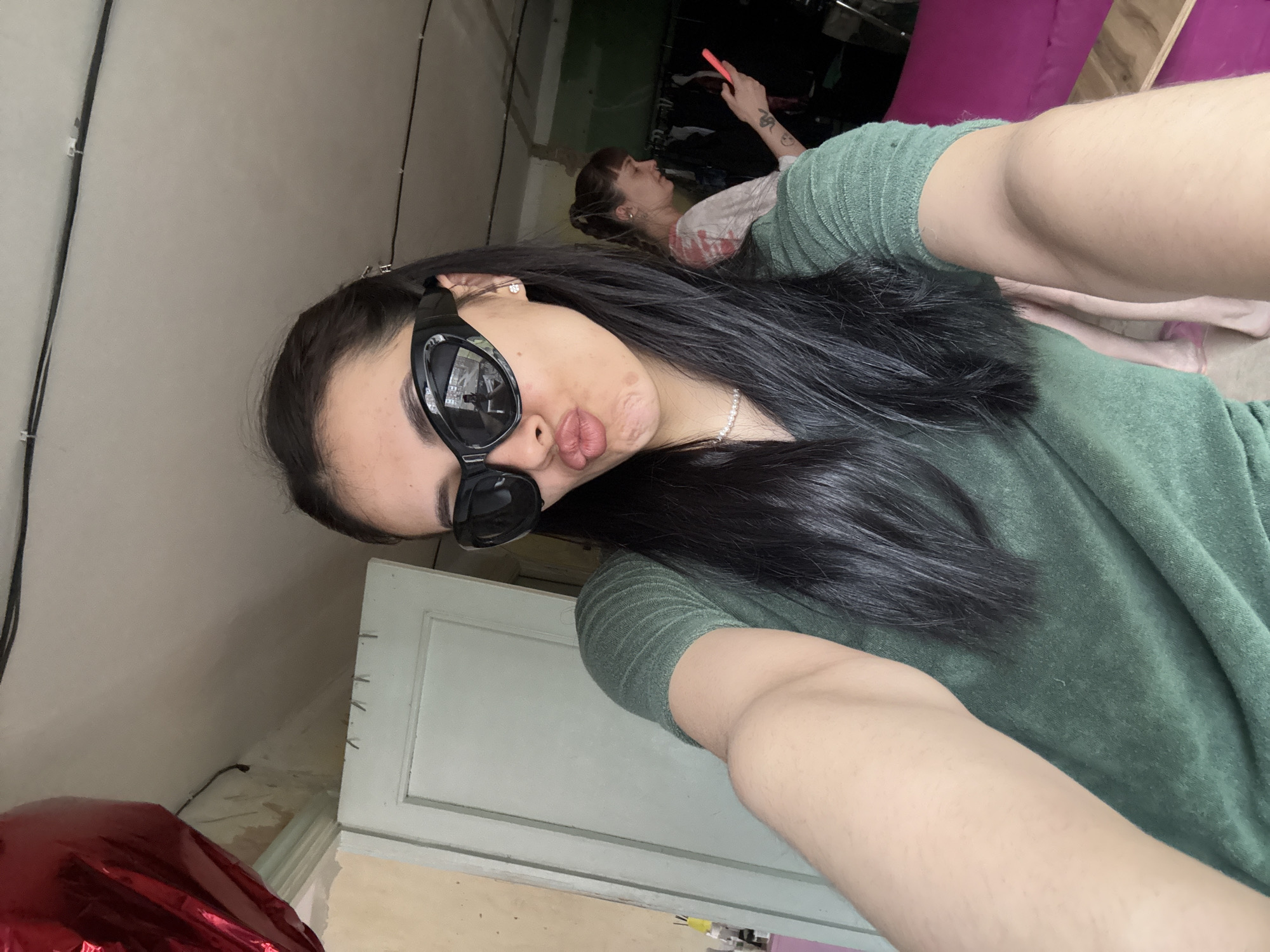 The image depicts a woman in the foreground, wearing sunglasses and a green top, posing for a selfie. Her hair is long and dark, and she's wearing a necklace. In the background, another person is lying down using a mobile phone. The setting appears to be indoors, possibly a room under renovation, with visible walls and what looks like furniture. No information in the image indicates the location to be Votkinsk, Russia, or any other specific city.