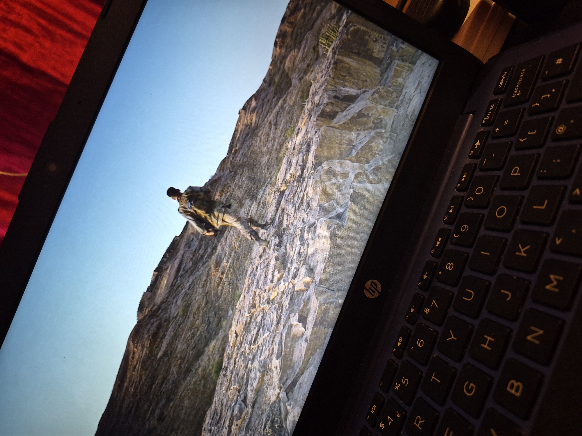 A close-up, angled shot shows a laptop screen displaying a scene of a person in military-style clothing walking across a rocky, mountainous terrain under a clear blue sky. The laptop is dark gray with a visible HP logo. Below the screen, the keyboard is visible, with keys labeled with letters, numbers, and symbols. The background is a blurred red surface. The image is taken from a low angle, looking up towards the screen and the person on it. There are no discernable landmarks or specific details that would indicate a location within the city of Sandwell, United Kingdom.