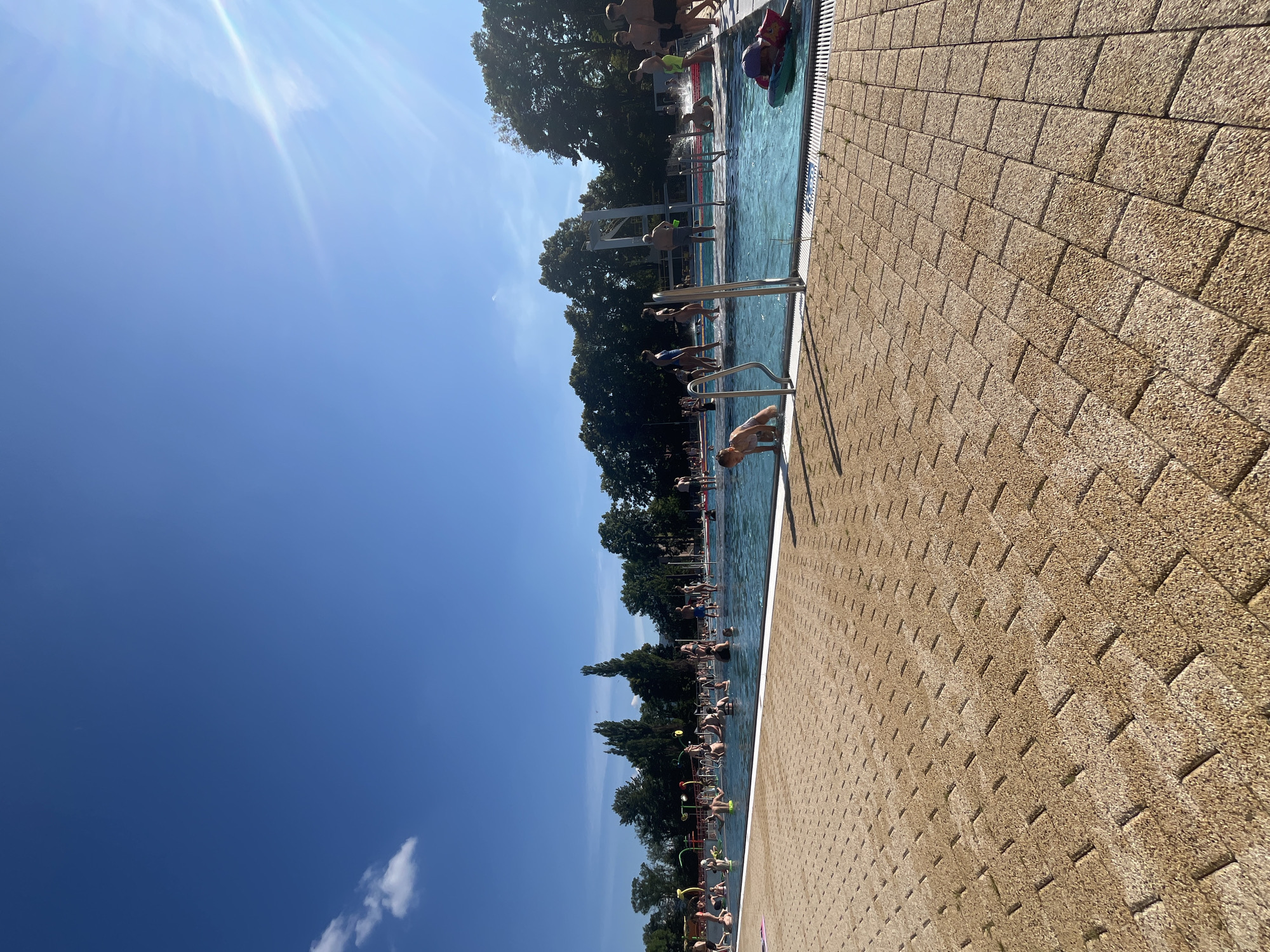 In an outdoor swimming pool on a clear day, many people are enjoying themselves. The pool has a shallow end with water jets and a deeper area. Some individuals are swimming, while others are wading or standing in the water. Children are playing and some adults are relaxing. The pool area is paved with light brown tiles, and lined with metal railings. In the background, lush green trees form a backdrop under a bright blue sky with a few wispy clouds. Sunlight streams into the scene, creating bright highlights and casting shadows.