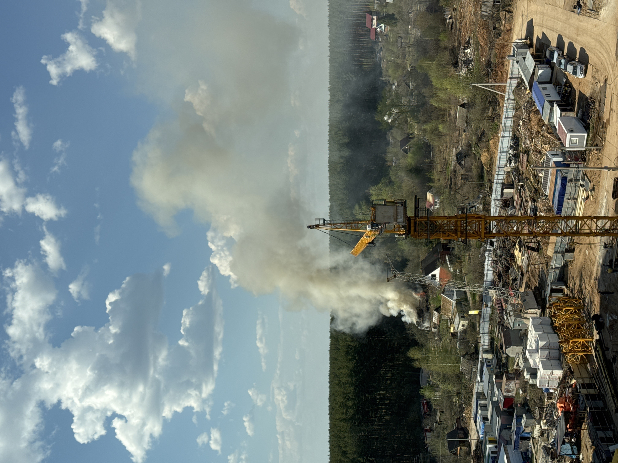 An elevated view of an incident occurring in Izhevsk, Russia. Visible from the elevated position are clouds in the sky, a forest line, and dense urban development. Smoke plumes rise from a fire, with a yellow construction crane in proximity to the fire. The area around the incident seems to be a mix of residential buildings and a construction site with building materials stacked neatly. Various objects such as trailers, vehicles, and construction equipment are on the right side of the frame, suggesting a staging area. The absence of first responders is noted in this view.