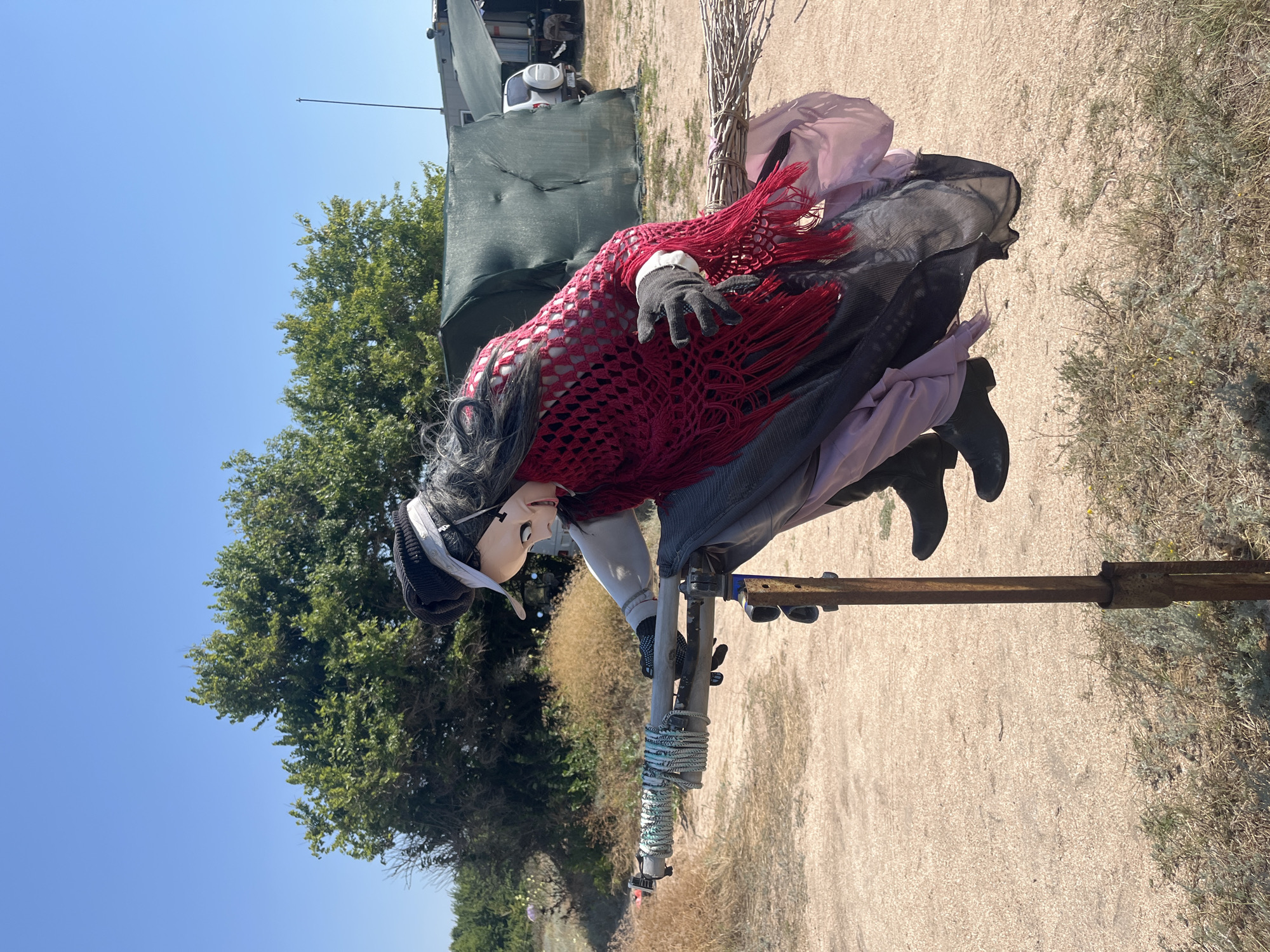 In Dolzhanskaya, Russia, a mannequin dressed as a witch is positioned outdoors, appearing to be riding a broomstick. The witch mannequin has long black hair, a pale mask-like face with a wide grin, a black hat, a white shirt, and a red crocheted shawl. It is wearing a long dark skirt and black boots. The broomstick is supported by a metal stand. The background shows a sandy ground, sparse vegetation, green trees, and some structures covered with tarps, under a clear blue sky.