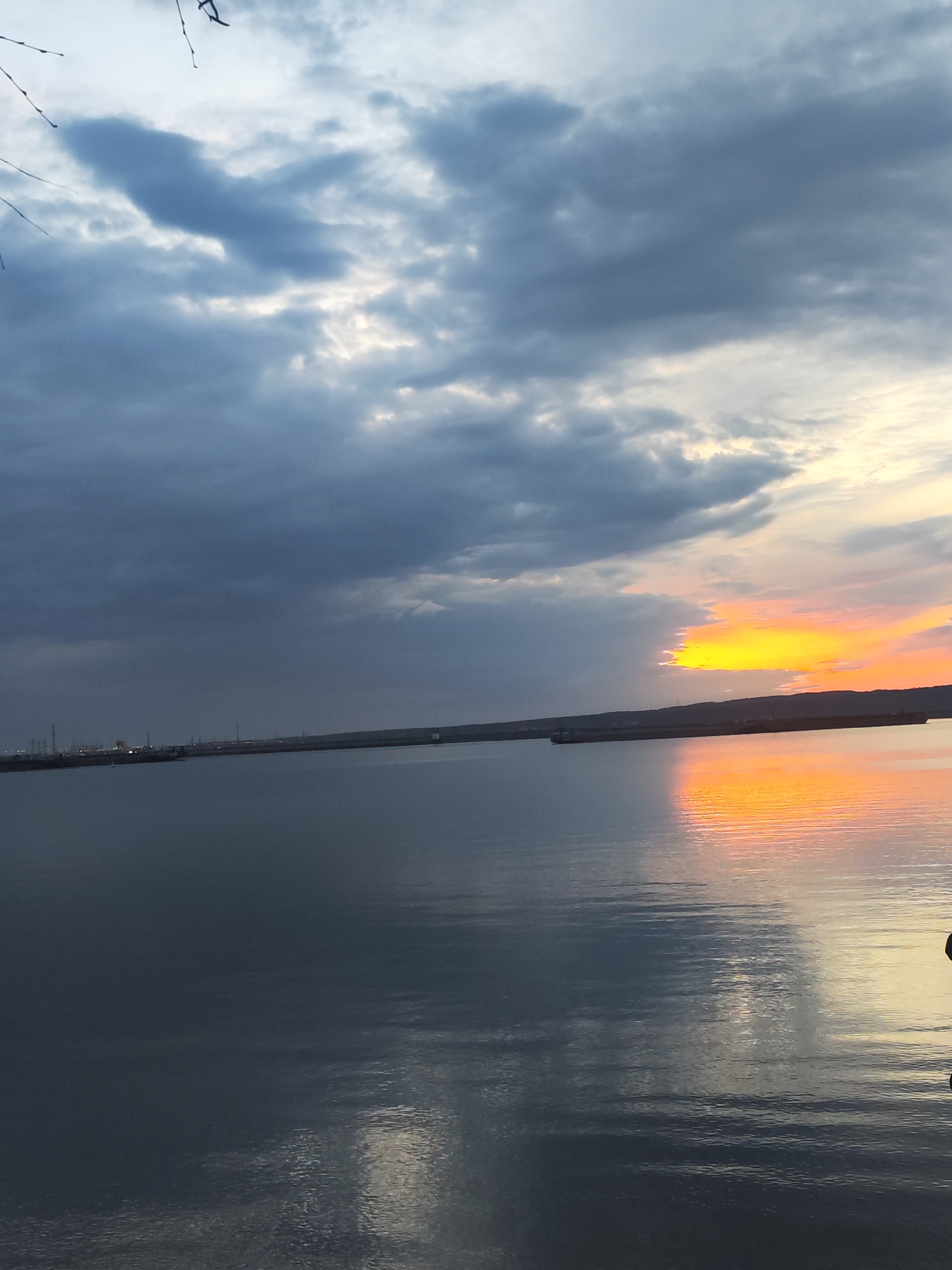 The image depicts a body of water reflecting the sky at sunset. Dark, cloudy skies contrast with a bright orange and yellow sunset on the right side of the image. In the distance, a dark, low-lying landmass stretches across the horizon. On the left, there are structures that appear to be part of an industrial area, and some sparse branches are visible at the top left corner. Based on the location provided, this is likely the Kama River near Chaikovsky, Russia. The scene is still and peaceful. There is no human activity immediately visible in the image.