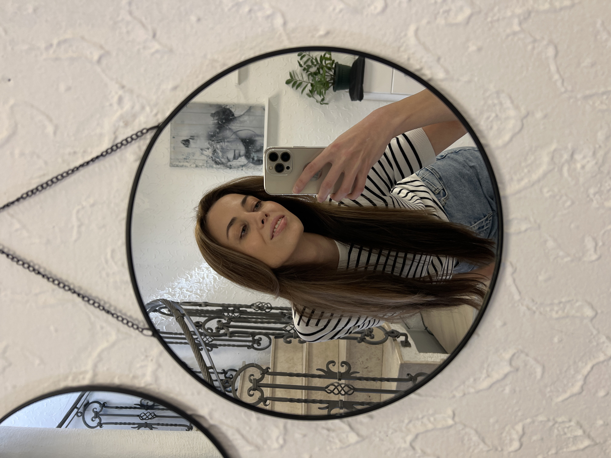 The image shows a reflection in a round mirror hanging on a textured white wall. A woman is taking a selfie in the mirror, holding a smartphone in her right hand. She has long brown hair and is wearing a striped top and jeans. Behind her, a staircase with a wrought iron railing is visible, as well as a painting and a potted plant on a shelf. The location is likely a residential interior in Bar, Montenegro, based on the style of the railing and architecture.
