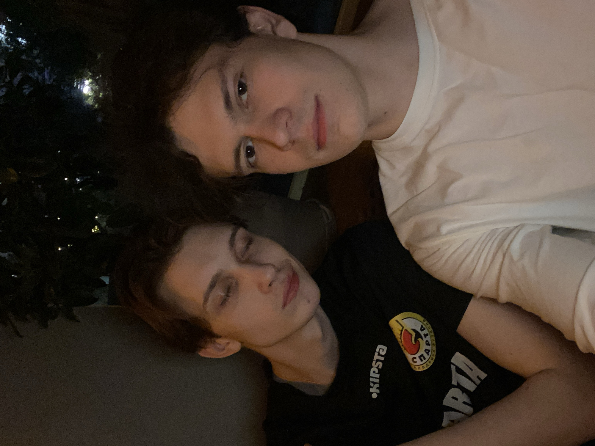Two young men are posing for a photo. One man, wearing a white shirt, is looking at the camera with a neutral expression. The other man, wearing a black shirt with a yellow and white logo featuring the word "SPARTA" on it and the brand name "KIPSTA", has his eyes closed and appears to be resting or asleep. The photo appears to be taken outdoors, with some greenery visible in the background. The location in the city of Krasnoyarsk, Russia, cannot be confirmed based on the image provided.
