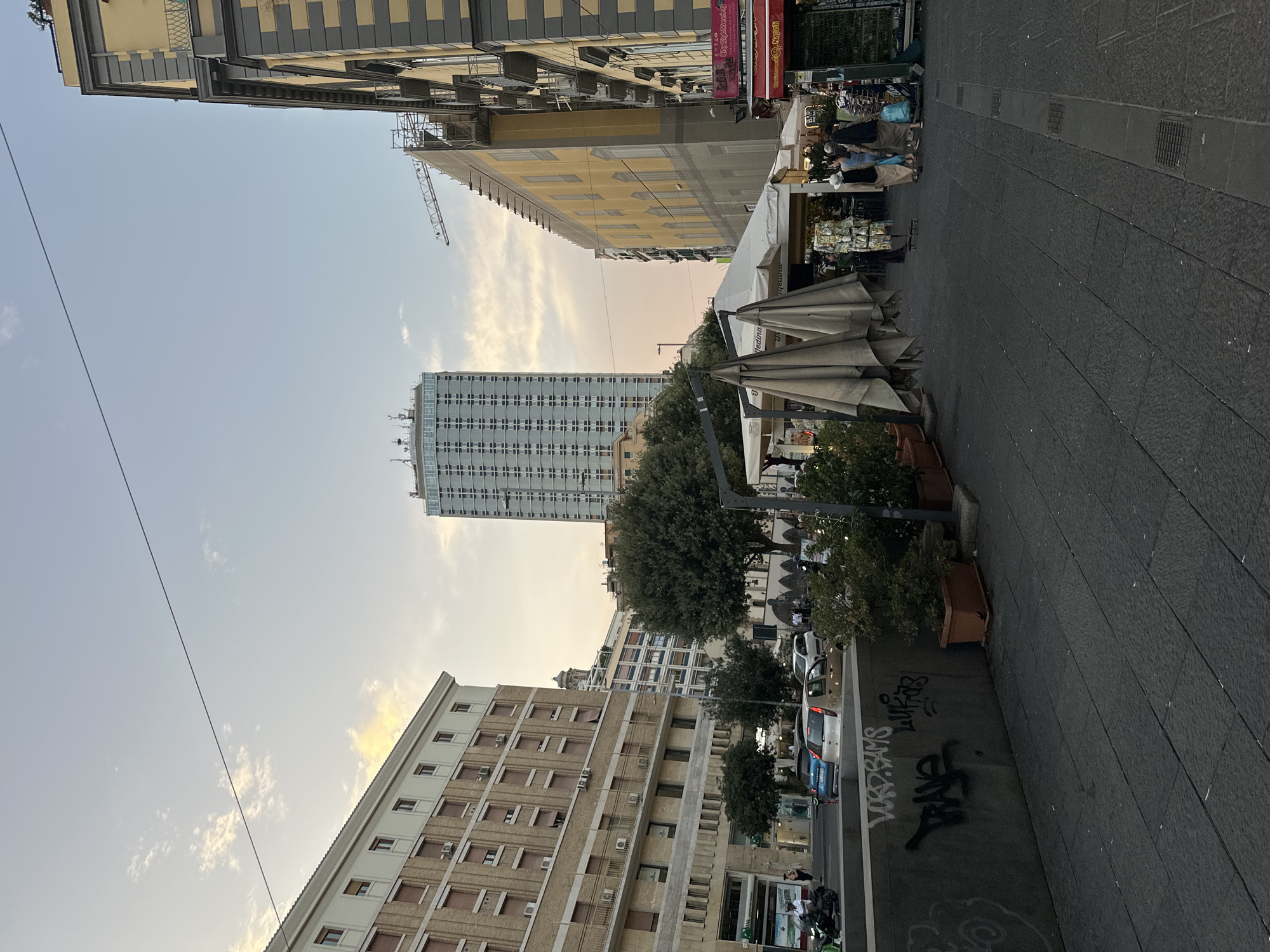 The image depicts a street scene in Naples, Italy, likely during the late afternoon or early evening given the soft lighting and sky color. On the left, a large, multi-story building with a light brown facade and numerous windows is visible, featuring several air conditioning units. Adjacent to it, at street level, is a storefront with awnings and signage. Further down the street, a tall, modern skyscraper with a grid-like facade dominates the skyline.
In the center of the image, a large tree stands prominently, with several smaller trees and potted plants in the foreground. To the right of the tree, a market stall with white tents and umbrellas is set up, displaying merchandise. A few people are browsing or walking past the stalls. Further to the right, another building with a yellow facade and grey architectural details is present, with some scaffolding suggesting ongoing construction or renovation.
The street in the foreground is paved with dark cobblestones. A low concrete barrier runs along the left side of the pedestrian walkway, covered in graffiti. Several cars are parked or driving on the street in the mid-ground, including a blue hatchback and a white sedan. The overall atmosphere is that of a busy urban environment, with a mix of historical and modern architecture.