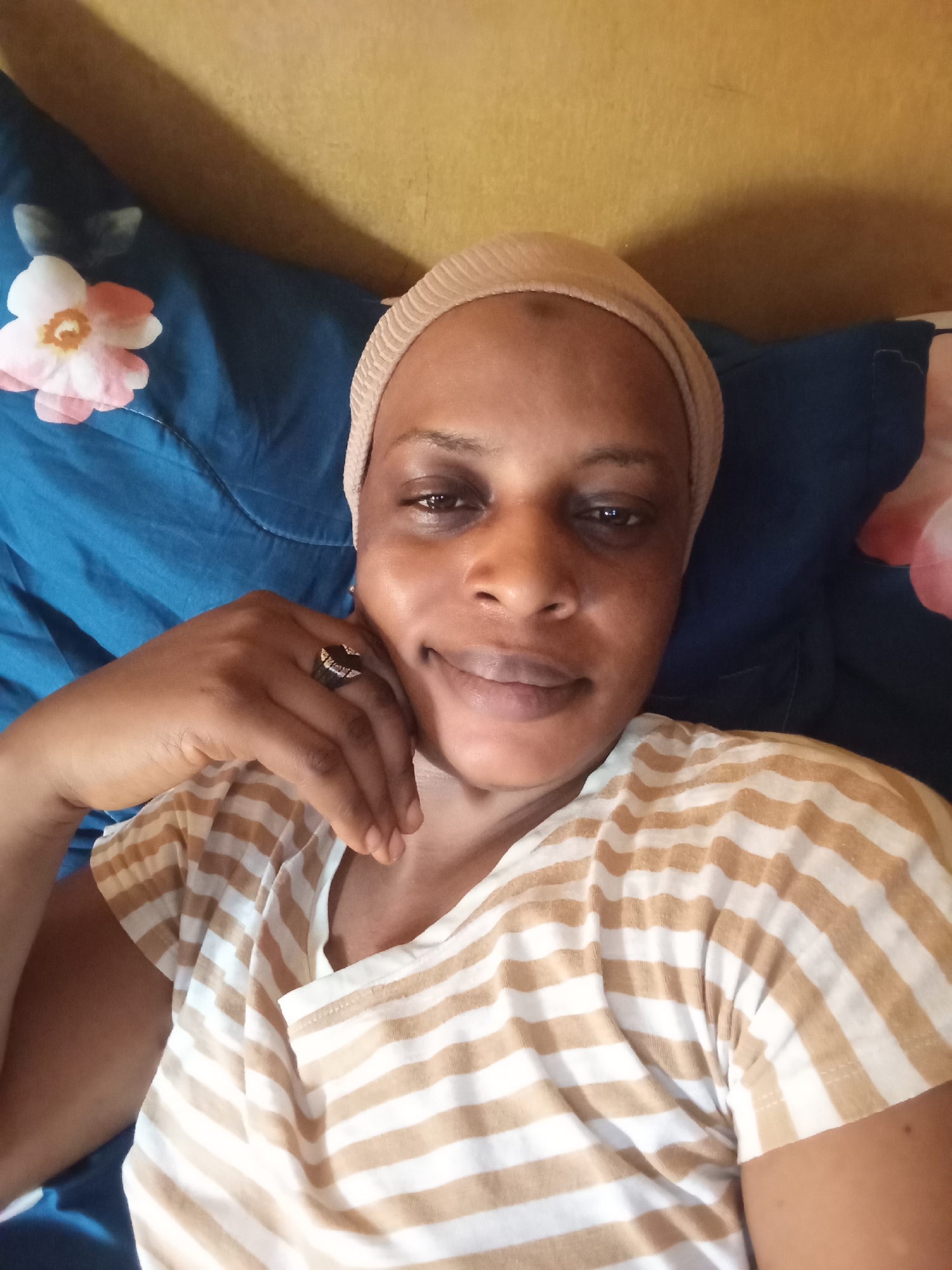 A medium shot depicts a person with brown skin lying on their side in what appears to be a bed. The person is wearing a tan head wrap and a tan and white striped t-shirt. Their left hand is raised and positioned under their chin. A dark-colored ring adorns their finger. The bedding consists of a blue pillow with pink and white floral patterns. A yellow wall is visible in the background. The location is presumed to be Narayi, Nigeria.