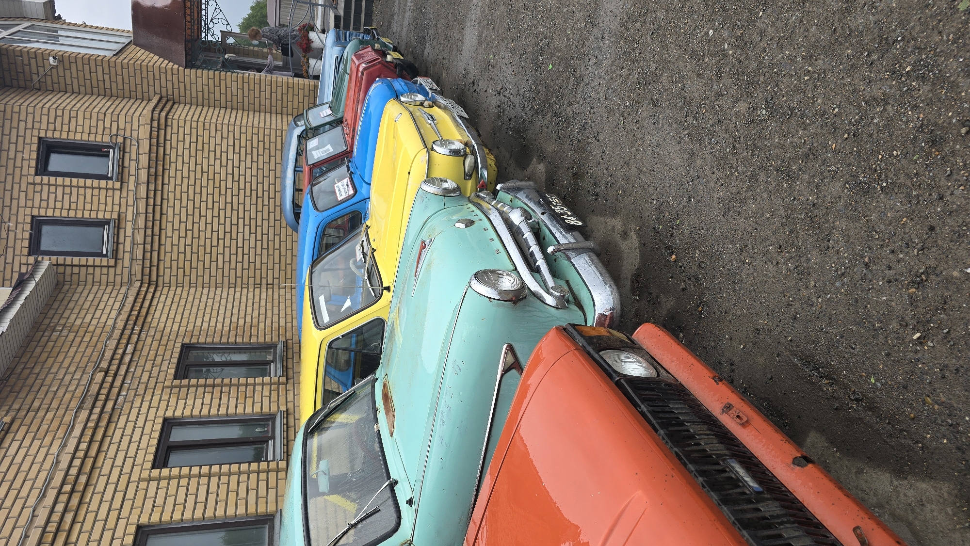 The image shows a line of vintage cars parked on a gravel surface next to a beige brick building. The cars are colorful, with a turquoise car in the foreground, followed by a yellow car, a blue car, and a red car further down the line. The building has several windows, and the ground appears wet, suggesting it might be raining or has recently rained. The sky is overcast, indicating a cloudy day. The scene is set in Bol'shoi Mashlyak, Russia. There is some visible text on the license plate of the turquoise car, which appears to read "BA 35-88 PB". No people are clearly visible in the image.