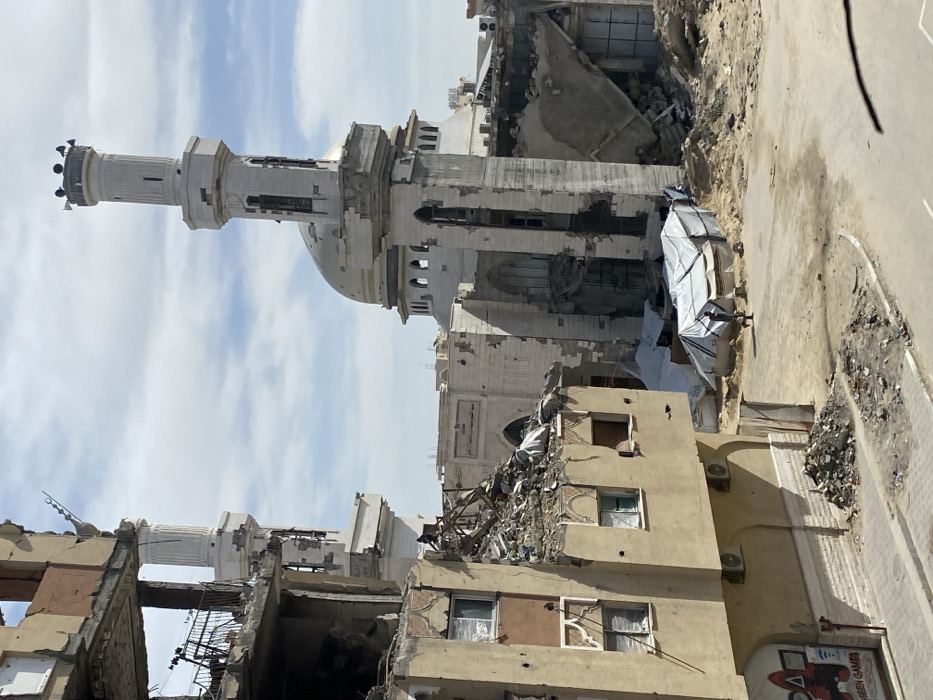 The image depicts an urban landscape in Khan Yunis, Palestinian Territory, characterized by extensive structural damage under a partly cloudy sky. In the background, the Al-Saniya Mosque is visible, featuring a large dome and two white minarets, one of which has multiple loudspeakers mounted at its peak. The mosque itself shows signs of significant damage, with visible sections of its facade and interior exposed. Arabic text reading "مسجد السنية الكبير" (The Grand Al-Saniya Mosque) is legible on one of its walls.
In the foreground and mid-ground, a multi-story building on the left is heavily damaged, with its upper floors partially collapsed and interiors exposed, revealing debris and structural elements. Some windows are covered with white material. On its ground level, a sign with red graphics and the text "ASEEN GAMES" is visible, along with two exterior air conditioning units. The area between this building and the mosque is strewn with vast amounts of rubble, sand, and debris, indicative of widespread destruction. Amidst this rubble, several makeshift structures covered with white tarpaulins are erected.
A single individual, dressed in dark clothing, is observed walking from left to right across the sandy, debris-laden ground near these temporary shelters. The overall scene presents a desolate environment marked by severe infrastructural damage.