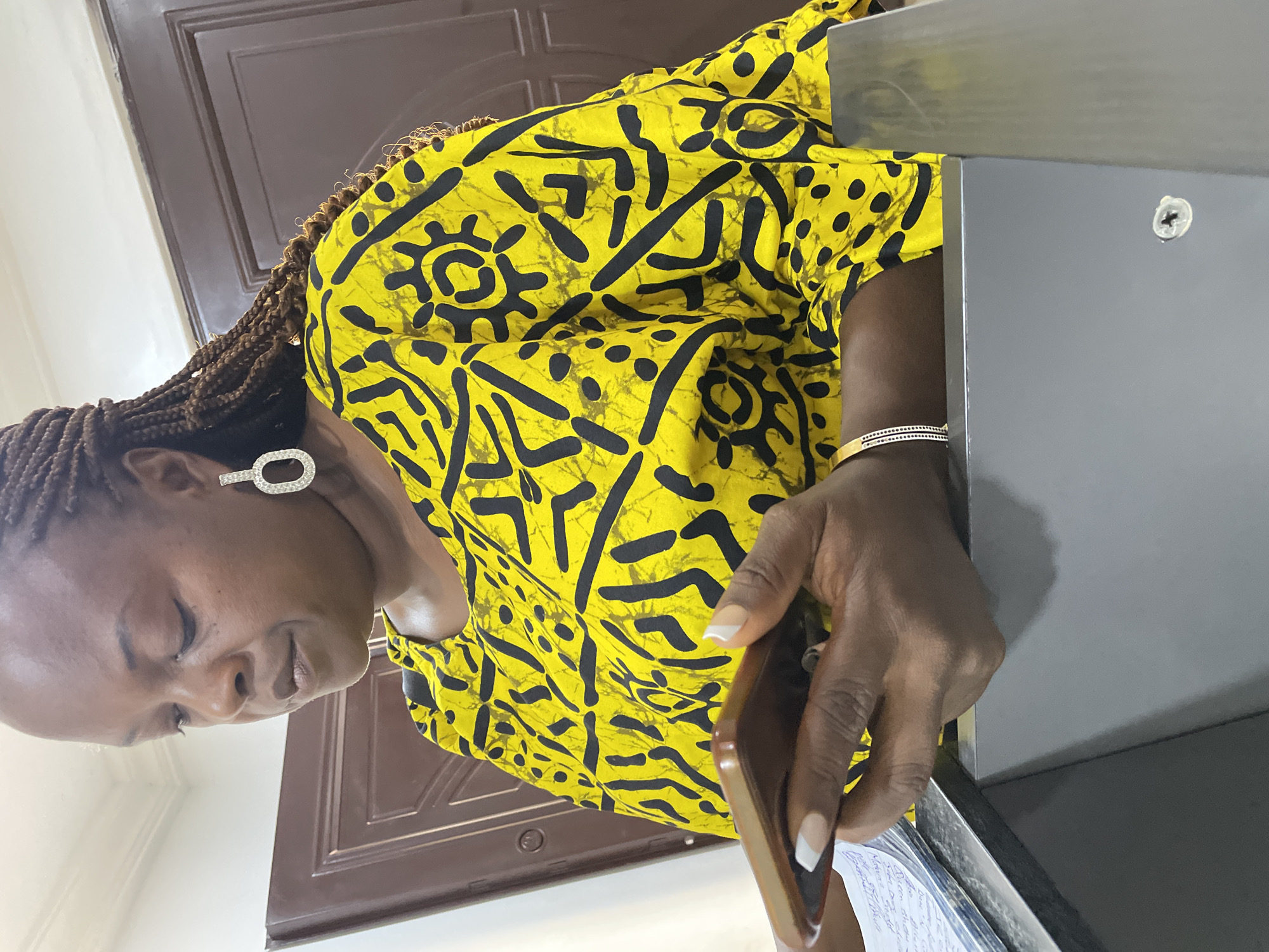 The image shows a dark-skinned woman with braided hair wearing a yellow and black patterned blouse. She is wearing an earring and a bracelet on her left wrist. She is looking down at a document and holding a brown object, possibly a phone. A gray metal object is on her right. A brown door is visible in the background. The location in the city of Jos, Nigeria is not discernible.