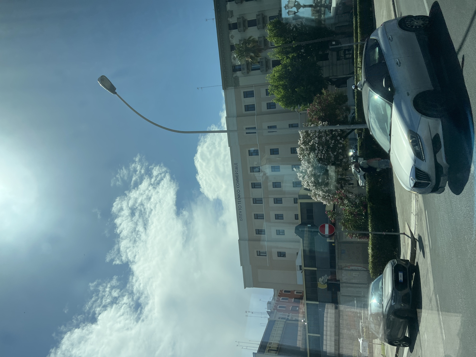 The image shows a street scene in Bari, Italy, with a focus on the "Istituto Tecnico Commerciale" building. In the foreground, two cars are parked on the side of the road. The car on the left is a dark gray Audi. The car on the right is a silver hatchback. A pedestrian, wearing shorts and a backpack, is visible near some flowering trees. The building itself is a multi-story structure with many windows. There is a street lamp arching over the road, and the sky is partly cloudy with bright sunlight. The street is paved, and there are trees and hedges lining the sidewalk. In the background, other buildings and architectural elements are visible, including balconies and palm trees.