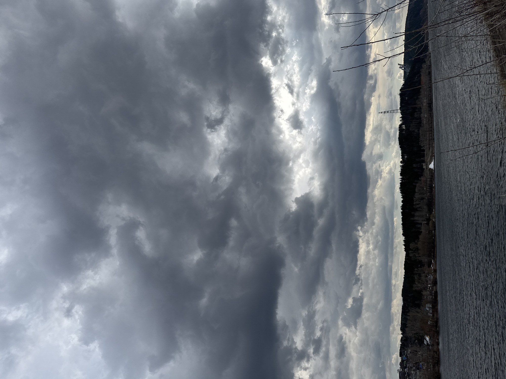 The image displays a dramatic landscape, oriented vertically, featuring a heavily clouded sky, a body of water, and a forested shoreline. The left and central portions of the frame are dominated by a vast expanse of dark gray, tumultuous clouds. Bright sunlight pierces through the cloud cover in vertical streaks, illuminating the edges and undersides of the clouds, creating a striking contrast and depth.
On the far right, a dark body of water, likely a lake or river, appears rippled, suggesting movement or wind. A dense line of bare, dark trees forms the shoreline, running vertically along the right side, separating the water from the sky and indicating late winter or early spring.
Visible within the forested area on the shoreline are two notable man-made structures: a tall, slender, white lattice transmission tower stands prominently, and further up the hill, a larger white structure with a dish-like antenna, possibly a radar station or observatory, can be seen. Small buildings or houses are also discernible along the base of the treeline. Thin, bare tree branches extend into the upper right corner of the frame.
The scene is captured during daytime, characterized by dynamic, overcast weather with intermittent sunbreaks. No people or visible text are present. The location context is Chaikovsky, Russia.