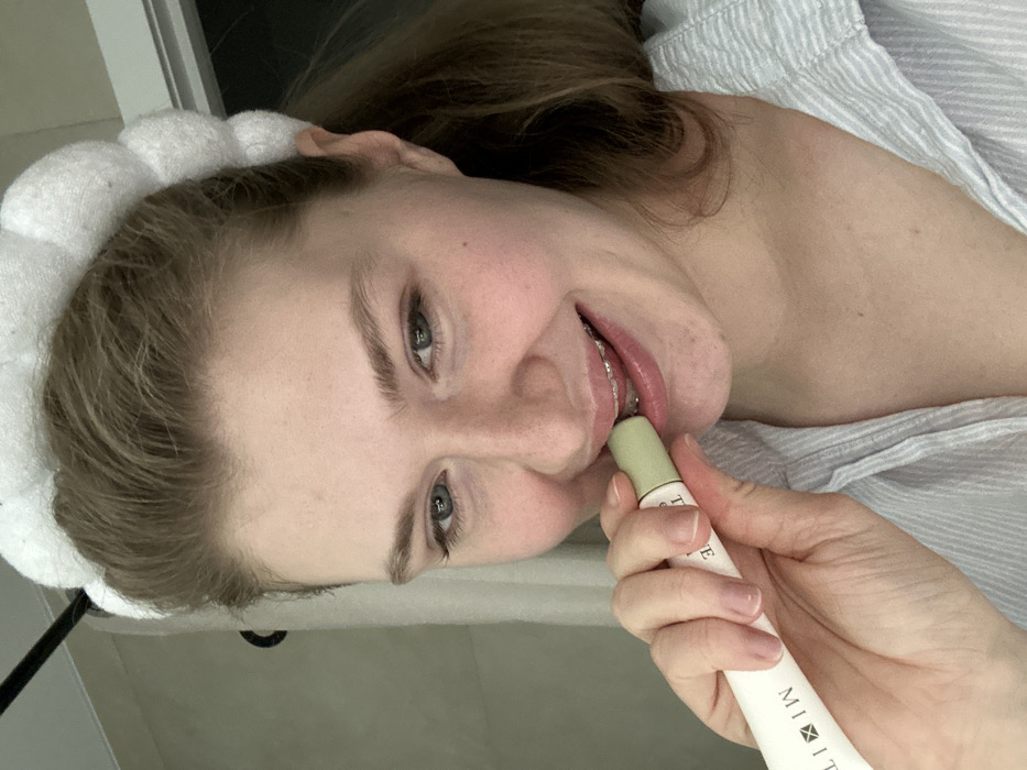 A person with fair skin, wearing a white, fluffy headband, applies lip balm. The person's head is tilted slightly to the side. Visible are the person's face, shoulder, and upper chest. The person holds a white tube of lip balm, with a green applicator. The balm has the text "MIIT" on it. The person has braces on their teeth. They wear a striped shirt. The background is a neutral color.