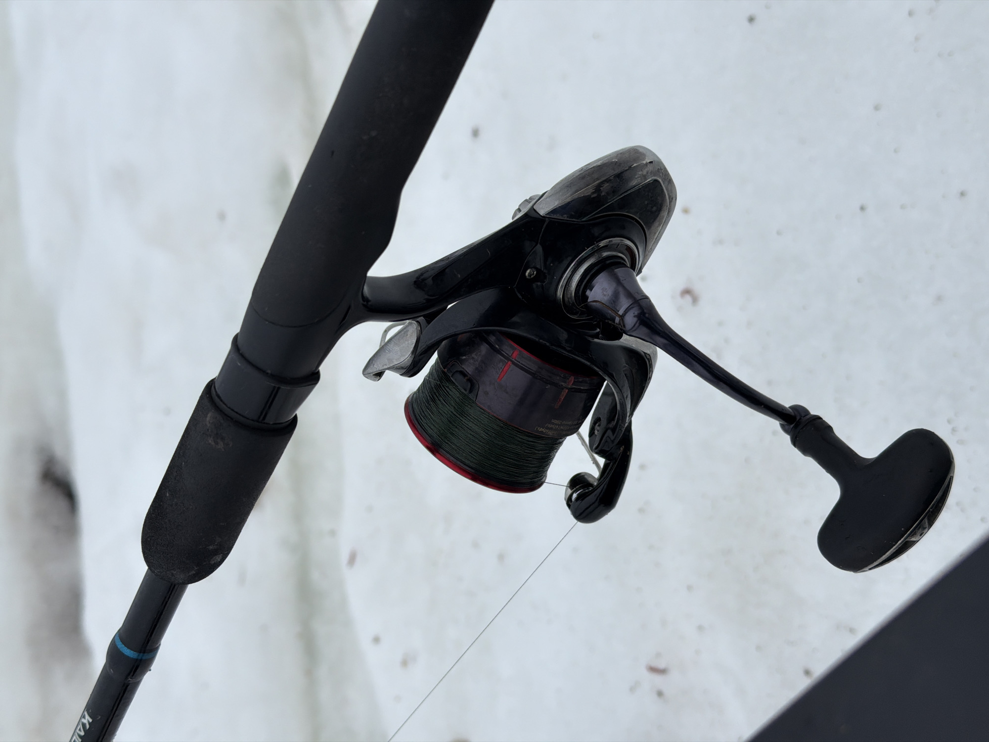 A close-up shot focuses on a fishing rod and reel against a snowy backdrop. The rod's grip is black and worn. The reel is attached to the rod, with a dark spool filled with green line. A fishing line extends from the reel. The environment shows patches of snow, with small, dark spots. The setting is plausibly outdoors. The location is assumed to be Chaikovsky, Russia.