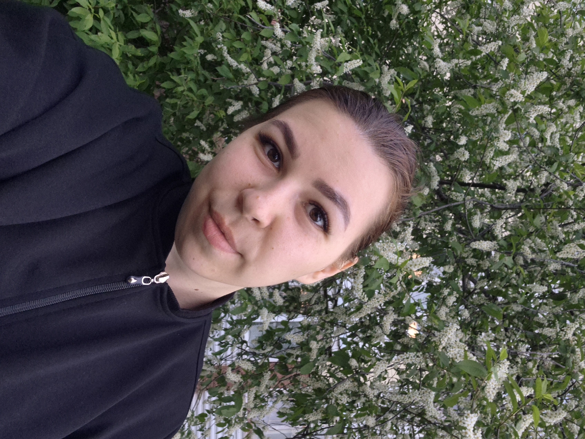 A woman with light skin, brown hair pulled back, and wearing a black zip-up jacket is taking a selfie. She is facing the camera at an angle, with her head tilted slightly. Behind her is a dense bush covered in small white flowers and green leaves, which suggests a park or garden setting. In the background, partially obscured by the foliage, is what appears to be a building with white siding and windows. The photograph was taken in Tobolsk, Russia.