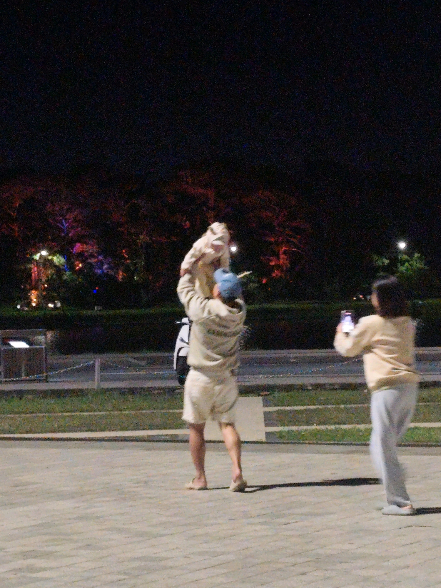 At night, in Da Lat, Vietnam, a person is holding up a baby or small child. The person is wearing a light-colored hooded sweatshirt and shorts. To the right, another person is standing and holding up a smartphone, seemingly taking a picture or recording video. The background shows trees illuminated with colorful lights, suggesting a festive or decorated area. A white scooter is visible behind the person holding the child. The ground is paved with rectangular stones.