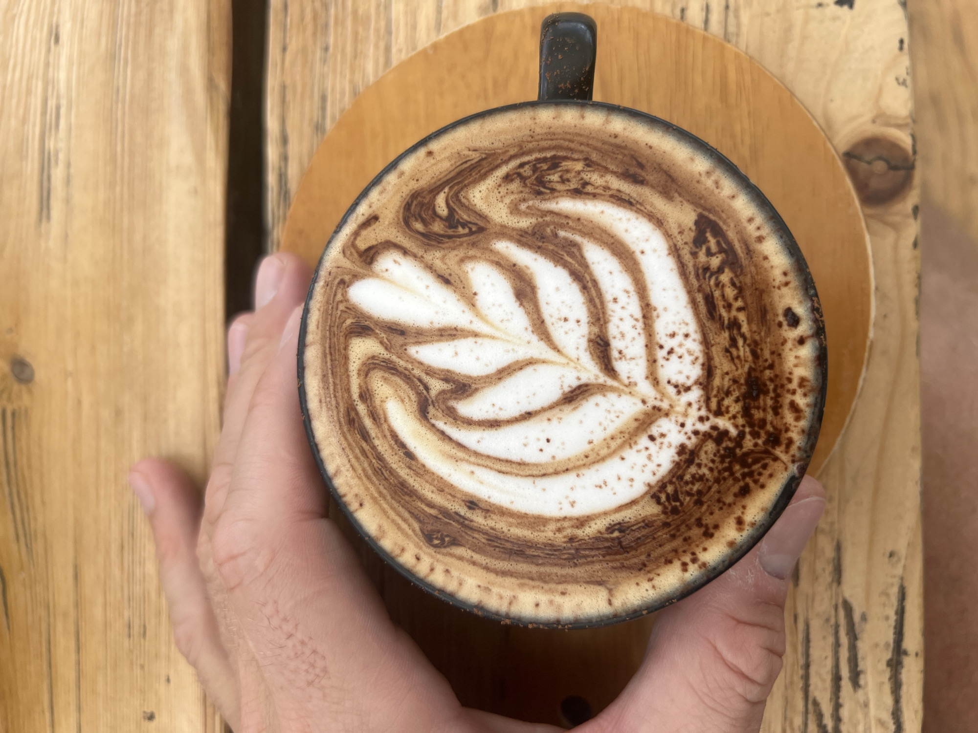 A high-angle shot captures a hand holding a black mug of latte art coffee, set on a circular wooden coaster. The coffee art features a leaf-like pattern in light foam against a dark brown coffee background. The mug and coaster sit on a wooden table made of light-colored planks. The image appears to be taken in the city of Baturiti, Indonesia.