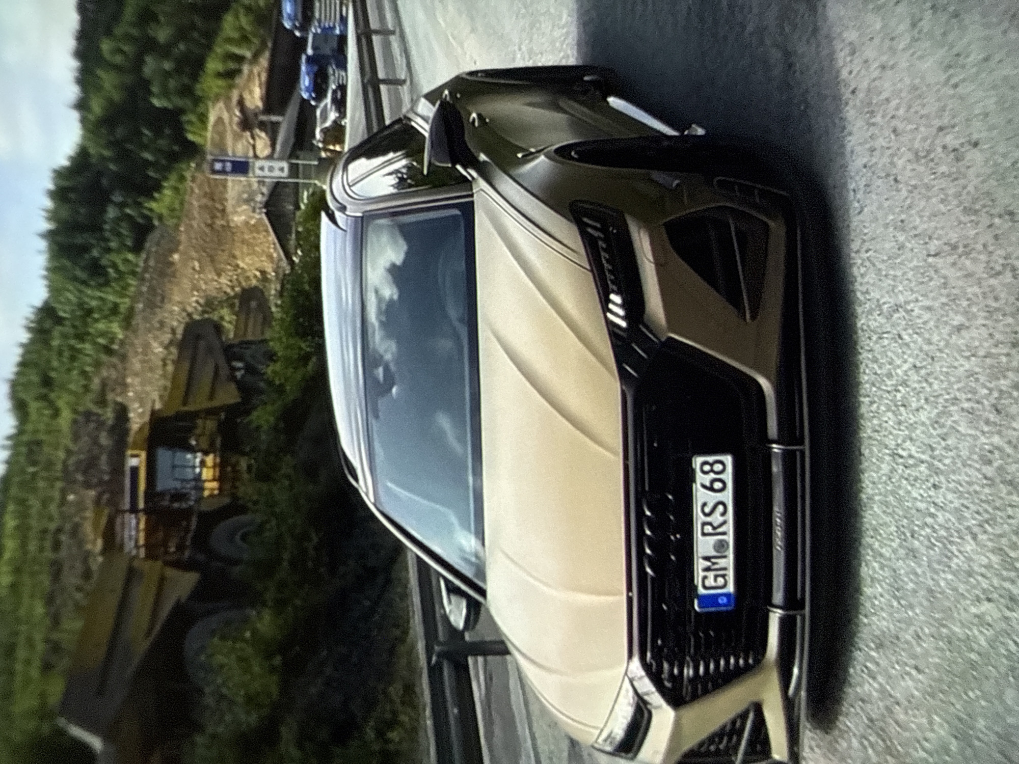 A matte gold Audi RS 6 Avant, featuring a black roof, dark grille, and black wheels, occupies the foreground, viewed from a front-right angle on a paved road. Its front license plate reads "GM RS 68", indicating a German registration from the Garmisch-Partenkirchen district. A dark grey guardrail runs along the left side of the road. In the background, a hilly landscape is visible, characterized by dense green trees and exposed, light brown earth and rock, suggesting a quarry or earthmoving site. A large yellow dump truck is partially visible on the left hillside. Towards the right background, a blue vertical sign stands near a building with a dark roof, with several semi-trailer trucks, including a blue one, visible on an adjacent roadway. The sky is partly cloudy. No people are present in the scene. The image provides no visual evidence to place the location in Varna, Bulgaria; the license plate explicitly points to Germany.