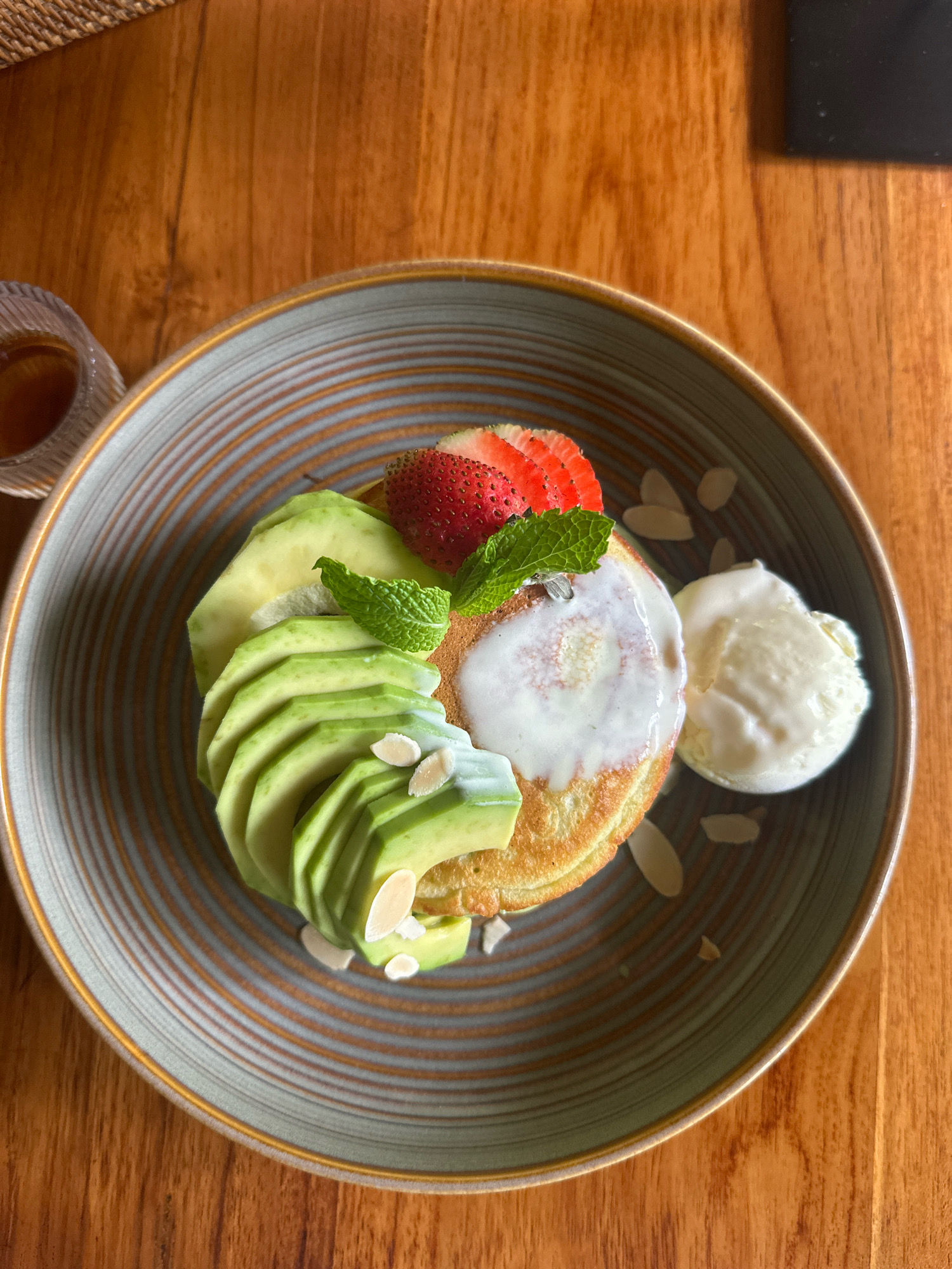 A close-up, overhead shot shows a plate of pancakes topped with avocado slices, a strawberry, mint leaves, and a dollop of yogurt, accompanied by a scoop of ice cream. Sliced almonds are sprinkled around the plate, which is set on a wooden table. A small glass of syrup is visible to the left. The setting appears to be a restaurant, as indicated by the presentation and the presence of other table items. The lighting is natural and comes from above, casting soft shadows. The image focuses on the food, with the background slightly blurred.