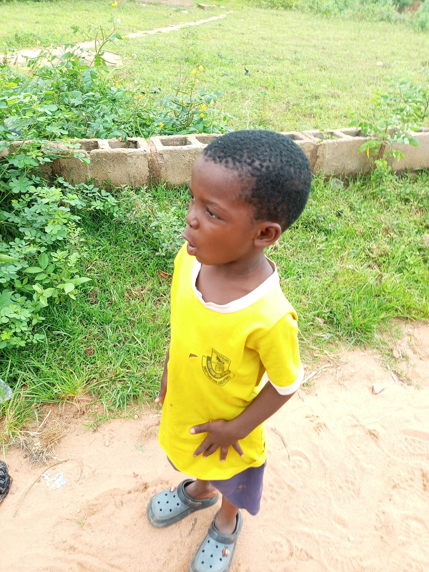 A young child stands on a dirt path beside a low wall made of cinder blocks. The child wears a yellow t-shirt with a circular emblem on the left side and blue shorts. Gray clogs are on the child's feet. Green vegetation, including leafy plants and grass, grows behind and to the left of the child. The background consists of more green grass. The scene is outdoors in what appears to be a rural or semi-rural area of Tempure, Nigeria.