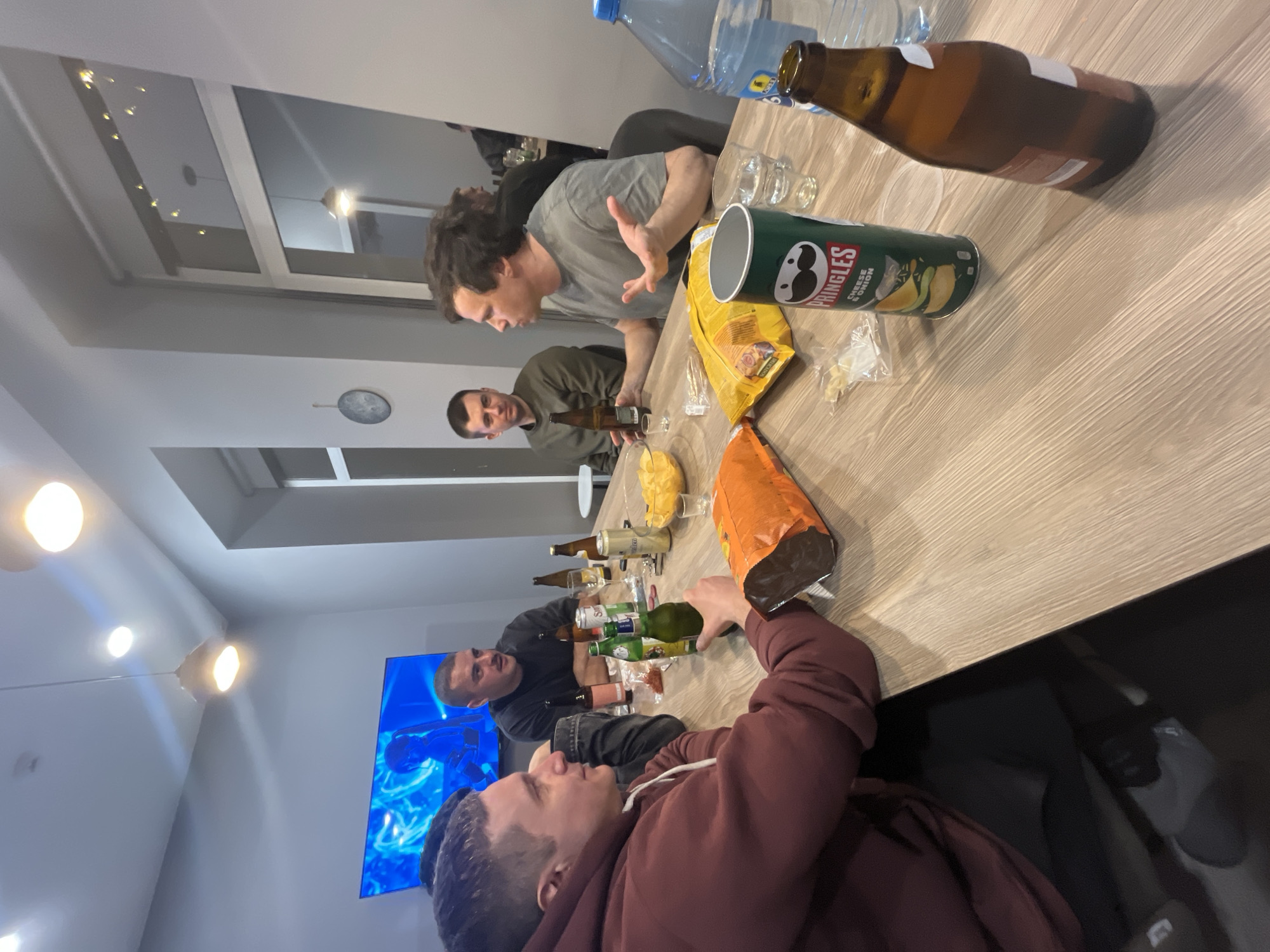 A group of four men are gathered around a light-colored wooden table indoors. There are open bags of chips and potato chips, several open beer bottles, and a large water bottle on the table. One man in a gray shirt gestures with his hands. Another man is opening a green beer bottle. A television screen displaying a blue and white image is visible in the background. The location is Unknown, Unknown.