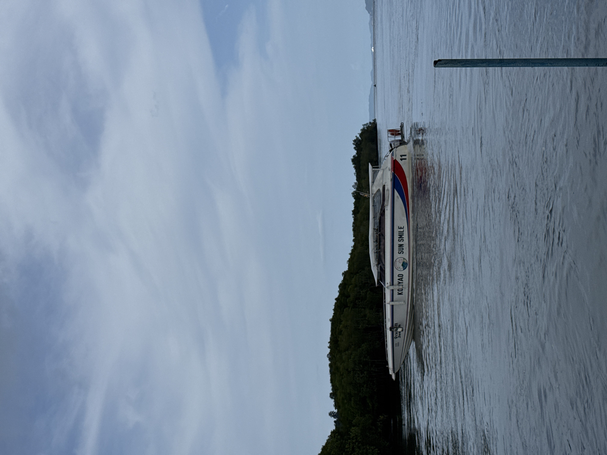 In Pa Khlok, Thailand, a white speedboat with red and blue stripes is moving through calm, grey water. The boat has "KO YAO" and "SUN SMILE" written on its side, along with the number "11". In the background, a dense green forest lines the shore under a cloudy sky. A blue pole sticks out of the water on the right side of the frame.