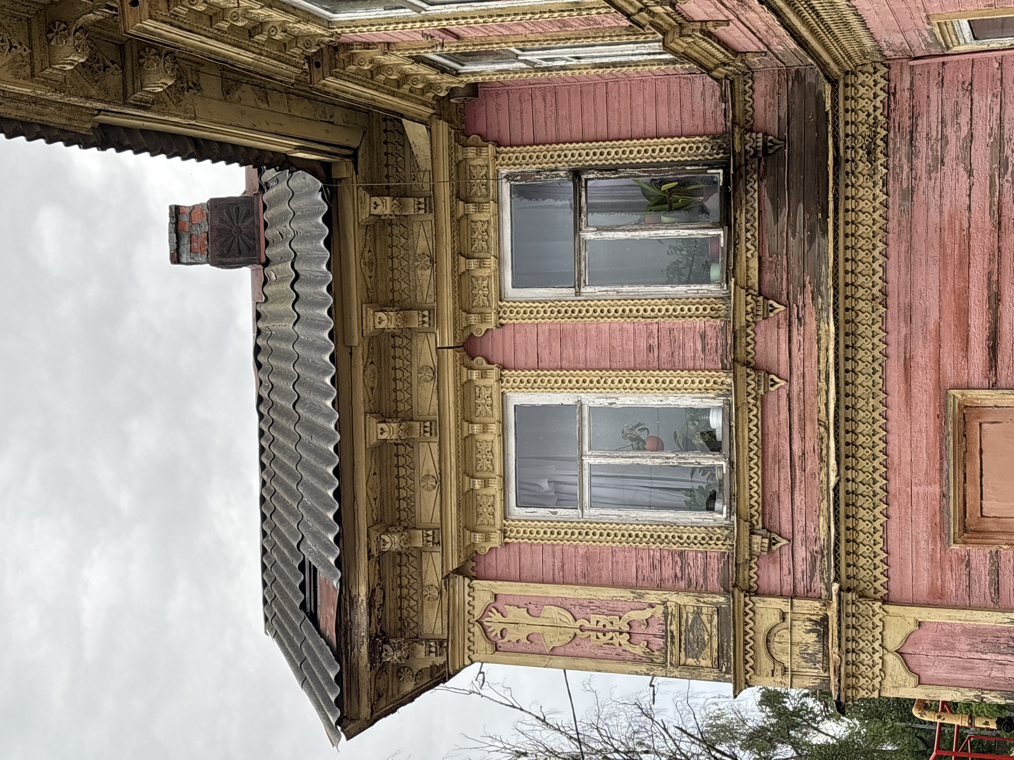 This is a close-up, eye-level exterior, medium shot of a weathered, historic wooden house with ornate yellow trim, located in Nizhny Novgorod, Russia. The house is painted pink, with visible signs of peeling paint and age. The upper portion of the house features a corrugated roof that appears to be in disrepair. Two windows with white frames are visible, each containing houseplants. Intricate wooden carvings adorn the eaves and window frames, showcasing a traditional architectural style. A brick chimney with a weathered cap is present on the roof. The sky is overcast and gray. The image does not contain any people.