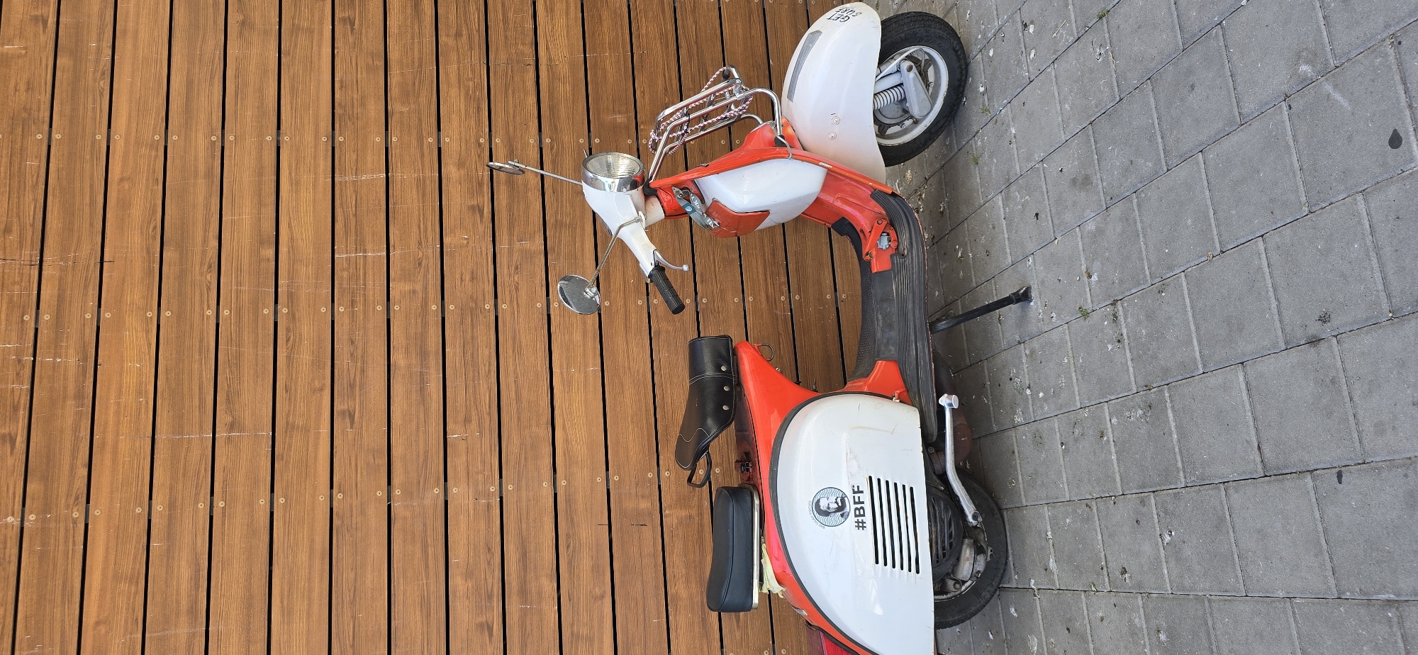 The image shows a vintage red and white scooter parked on a paved walkway next to a wooden deck. The scooter has chrome accents, including a luggage rack on the back and mirrors. A black seat is visible on the scooter. On the side of the white panel of the scooter, a circular sticker with a black and white image and text is present. The text on the sticker reads "#BFF". The wooden deck appears to be made of planks with visible screws. The paved walkway is made of gray rectangular bricks, some of which are marked with bird droppings. The image is taken from a high angle, looking down at the scooter. The lighting suggests it is daytime. The scene appears to be outdoors, likely in a public area or a residential yard. There is no specific indication of weather conditions, but the lighting suggests it is not overcast. The overall impression is of a static scene, with the scooter being the primary subject.