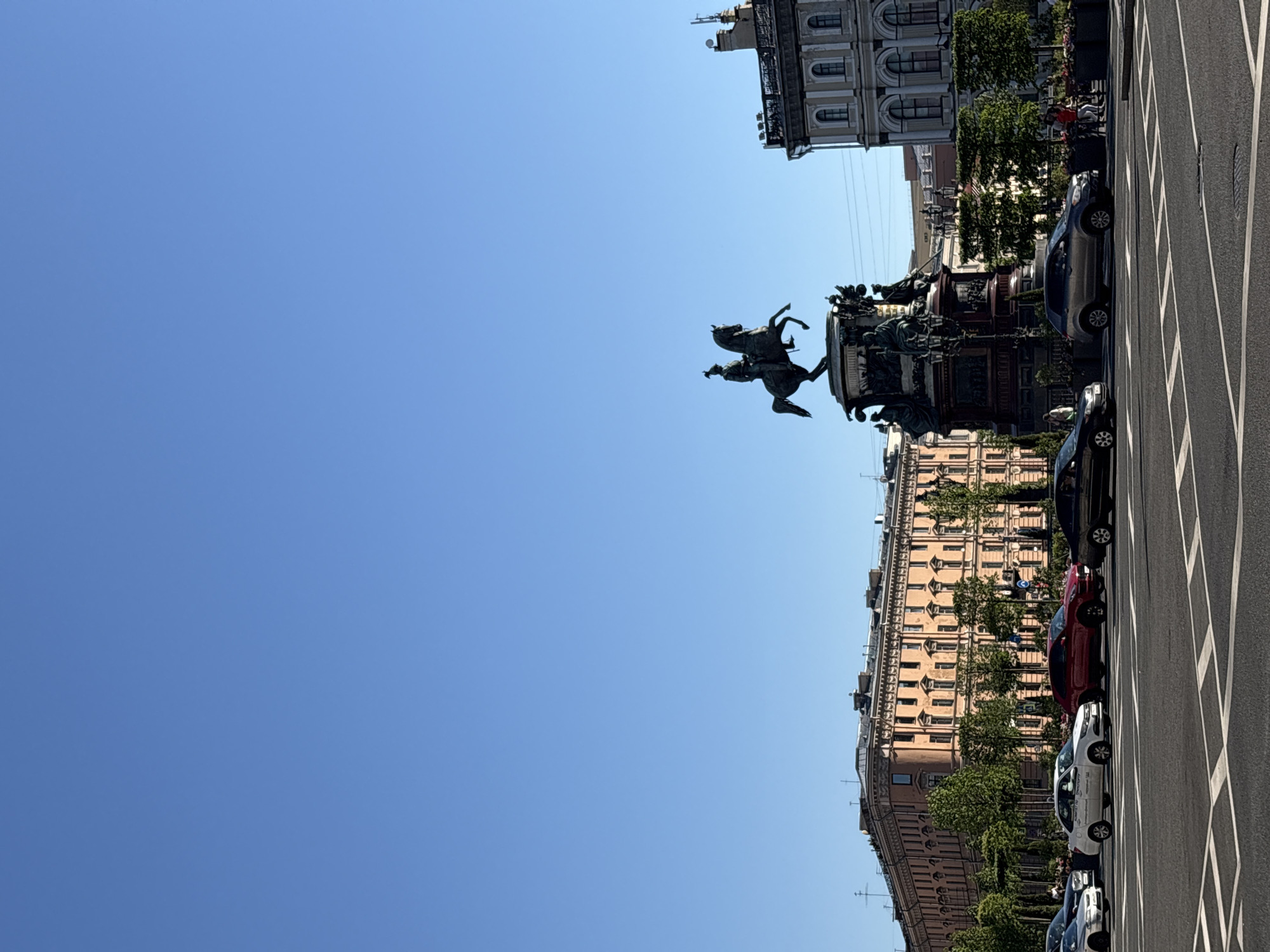 This image captures a street scene in Saint Petersburg, Russia, featuring the Monument to Nicholas I in Saint Isaac's Square. The upper half of the frame is dominated by a clear, bright blue sky devoid of clouds. Below the horizon, an asphalt road occupies the foreground, marked with white painted crosswalk lines and lane indicators.
A line of several cars is visible on the road. From left to right, these include a silver sedan, a white sedan, a red SUV, a dark grey SUV, and a black sedan. Another dark grey SUV is positioned closer to the camera, partially obscuring the monument.
The Monument to Nicholas I, an equestrian bronze statue, stands on a tall, intricately decorated pedestal in the mid-right portion of the image. The statue depicts a figure in military attire astride a rearing horse. The multi-tiered pedestal features various bronze figures and relief panels.
Behind the cars and monument, two multi-story buildings are visible. To the left, a long, light-orange building with classical architectural details, including numerous windows and decorative cornices, stretches horizontally. Green deciduous trees are planted along the street in front of this building. To the right of the monument, a light-grey building with arched windows on its lower visible floors and a roof adorned with antennas and chimneys is partially captured. Several indistinct human figures are visible on the sidewalks near the monument and buildings, appearing to be walking or standing.