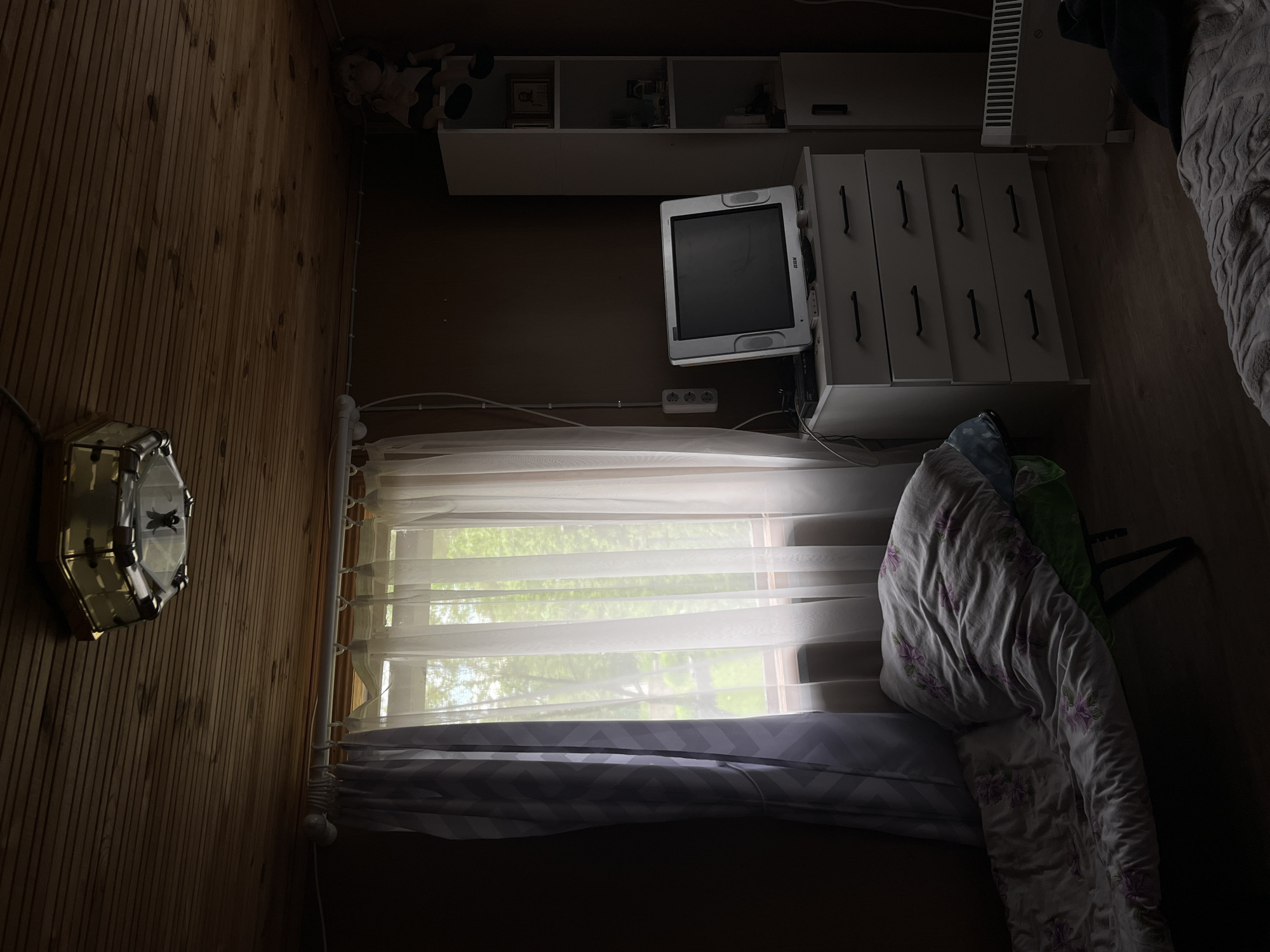 The image depicts an interior scene, likely a bedroom, in Мшинская, Russia. The room has wood-paneled walls on the left and a brown wall towards the top. A light fixture is mounted on the left wall. A window with white, sheer curtains is in the center. Outside the window, foliage can be seen. Above the window are wall-mounted shelves and a white cabinet with drawers. A CRT television sits on top of the cabinet. A bed covered with a floral-patterned comforter is on the right. A radiator is visible above the cabinet, partially obscured by a dark object.