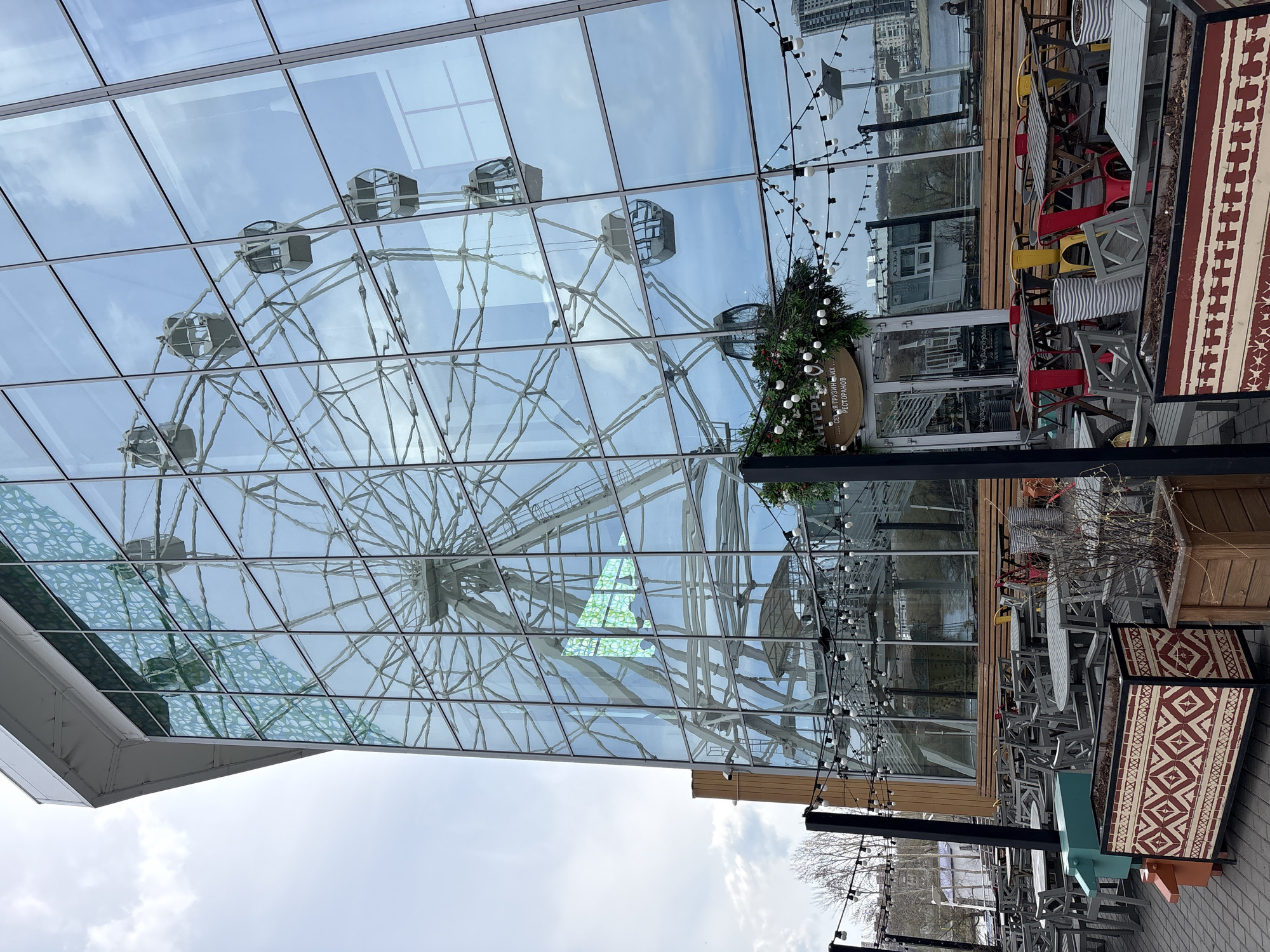 The image shows a reflection of a Ferris wheel in the glass facade of a building. Outside seating with tables and chairs suggest a restaurant or cafe nearby. String lights are strung across the outdoor dining area. The location is possibly in Ulyanovsk, Russia, but that cannot be verified from this image. The scene is still and shows no specific activities taking place.