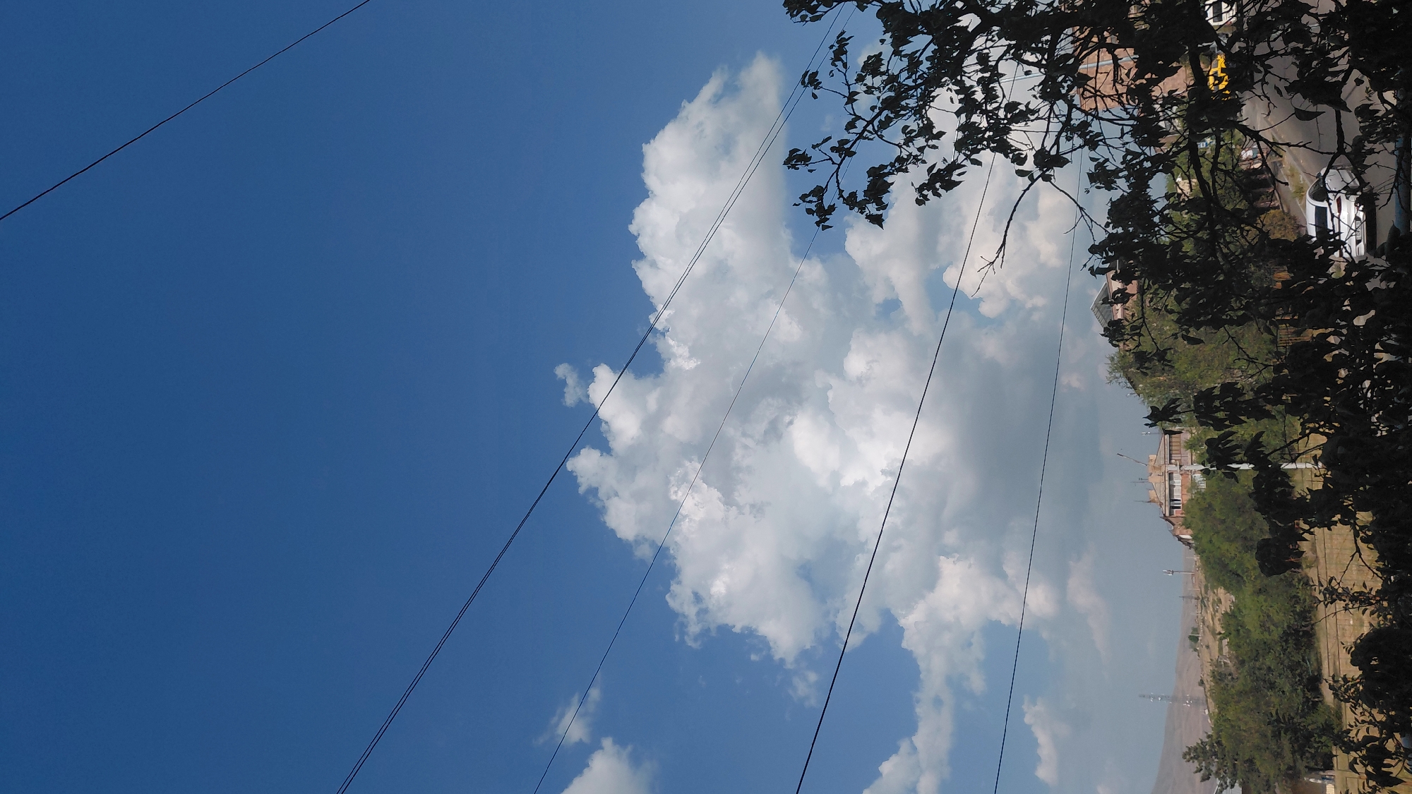 The image captures a street scene in Abovyan, Armenia, under a bright blue sky with scattered white clouds. Overhead, multiple dark electrical wires stretch across the frame, intersected by a silhouetted tree with dense foliage in the foreground. In the middle ground, a road curves uphill, with several buildings and lush green trees lining its sides. A white car is visible on the road, as well as a yellow vehicle further up. Distant hills form the backdrop, with some structures and communication towers on their slopes. The overall impression is that of a sunny day in a residential area.