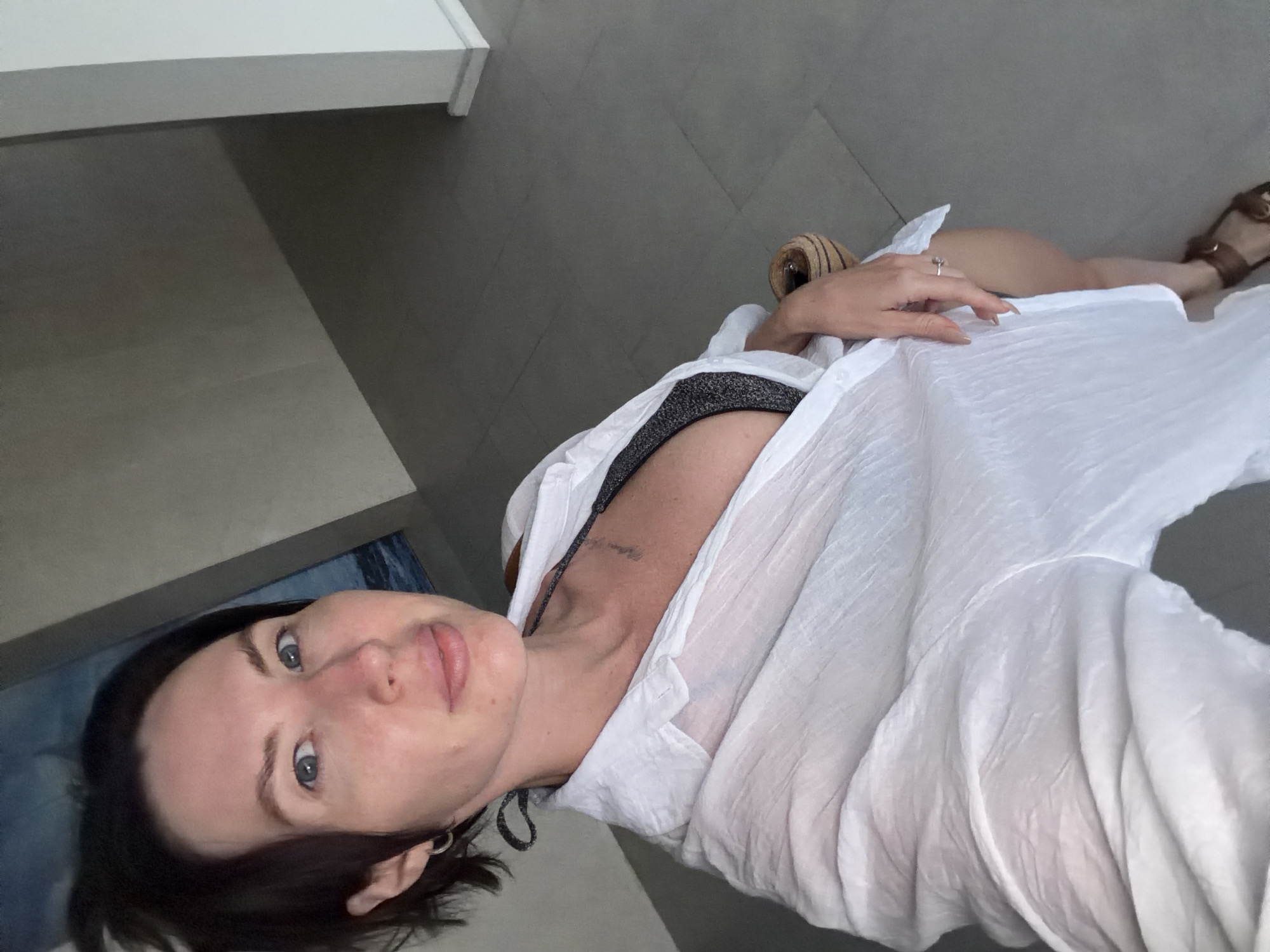 A woman with short dark hair and light skin, wearing a white unbuttoned shirt over a dark bikini top and a ring on her finger, is captured in a selfie taken from a high angle. She is sitting in a chair, partially visible on the right side of the frame. Her gaze is directed towards the camera. The background consists of a tiled floor and a wall, with a framed artwork depicting a seascape on the wall behind her. The image was taken in Dong Ninh Hoa Ward, Vietnam.