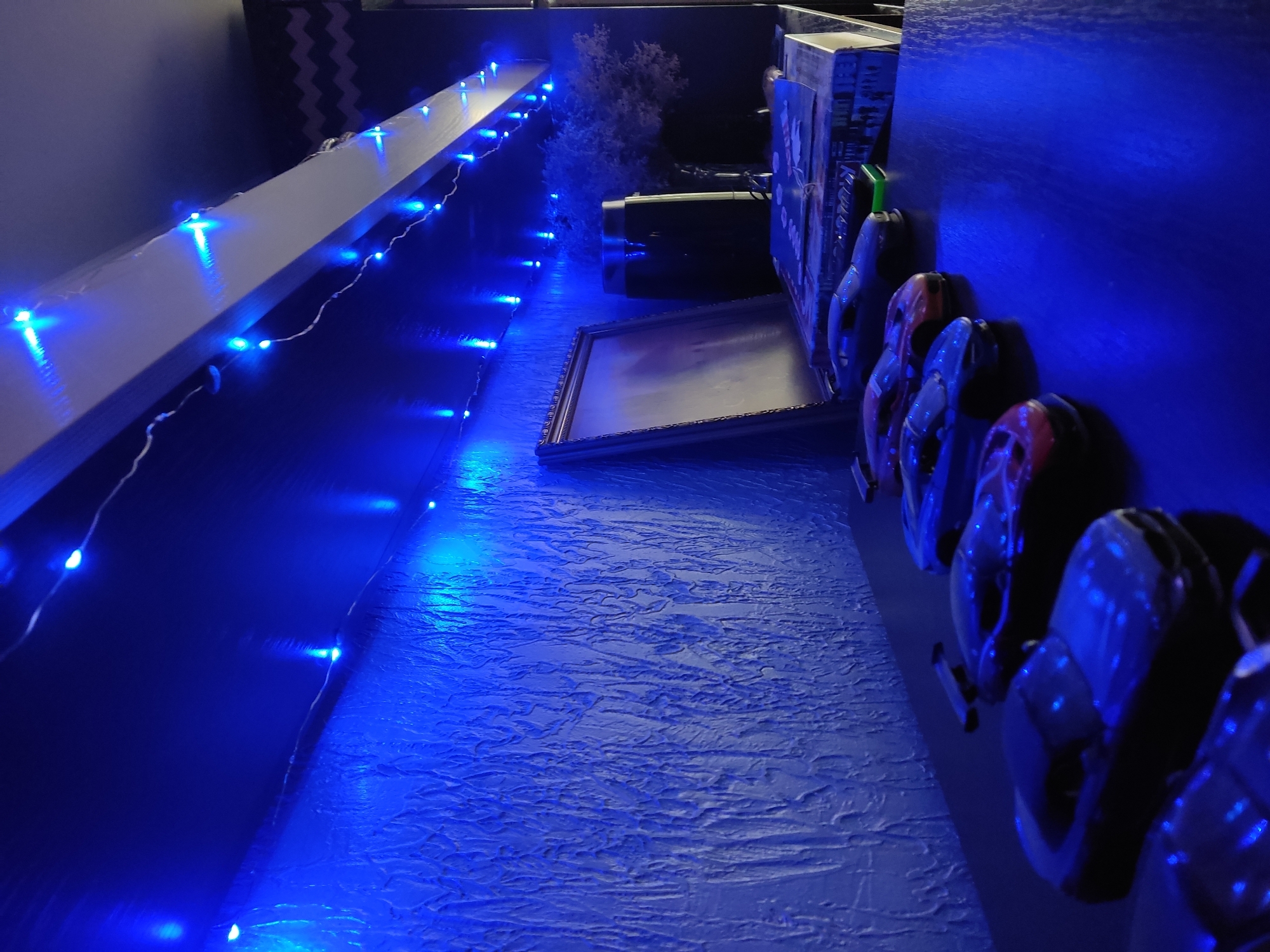 The image shows a balcony at night, illuminated by blue string lights. The balcony wall is textured and painted blue. On the right side of the image, several pairs of headphones are hung along the wall. Further back, a picture frame is leaning against the wall. In the background, a small potted plant is visible. Based on the visible objects and architectural features, the location could potentially be in Krasnodar, Russia. However, there is no definitive information in the image to confirm this location.
