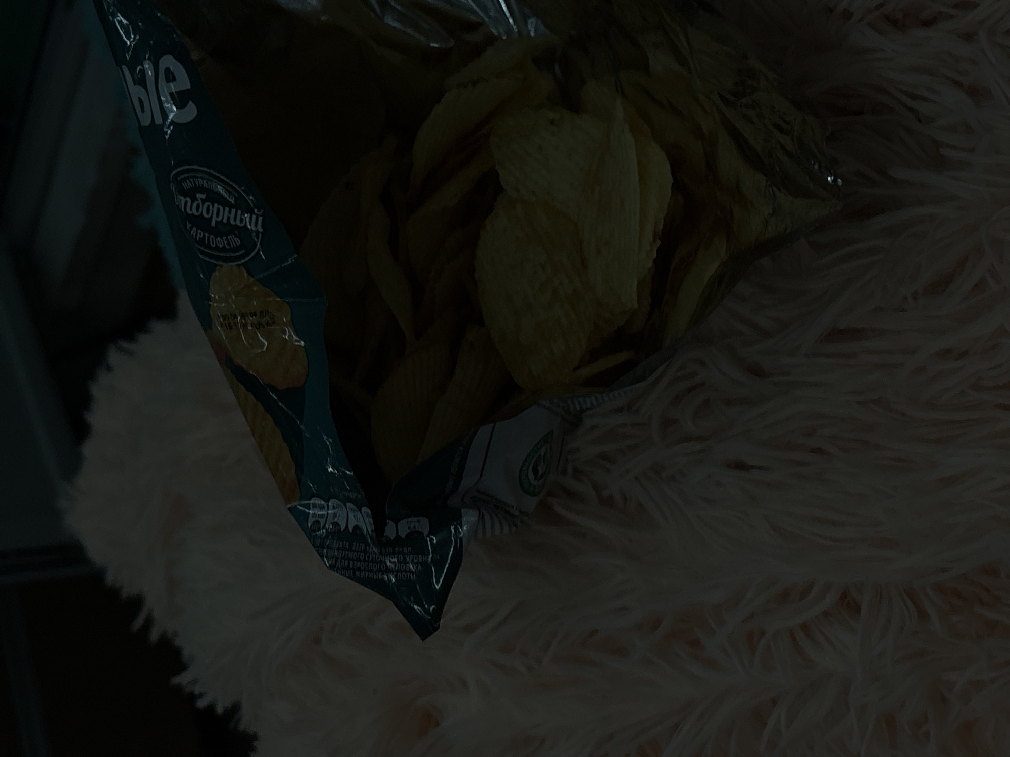 A bag of chips is shown spilling onto a fluffy, cream-colored surface. The bag is partially open, revealing the potato chips inside. The bag has a teal and silver design with text in Russian, including the word "Сырое" which translates to "raw" or "crude" and "КАРТОФЕЛЬ" meaning "potato". The scene is dark with soft lighting, suggesting it may be indoors. There are no people visible in the image. The location in Khimki, Russia, is not discernable from the image itself, as it is a general indoor scene with no specific landmarks or identifying features of the city.
