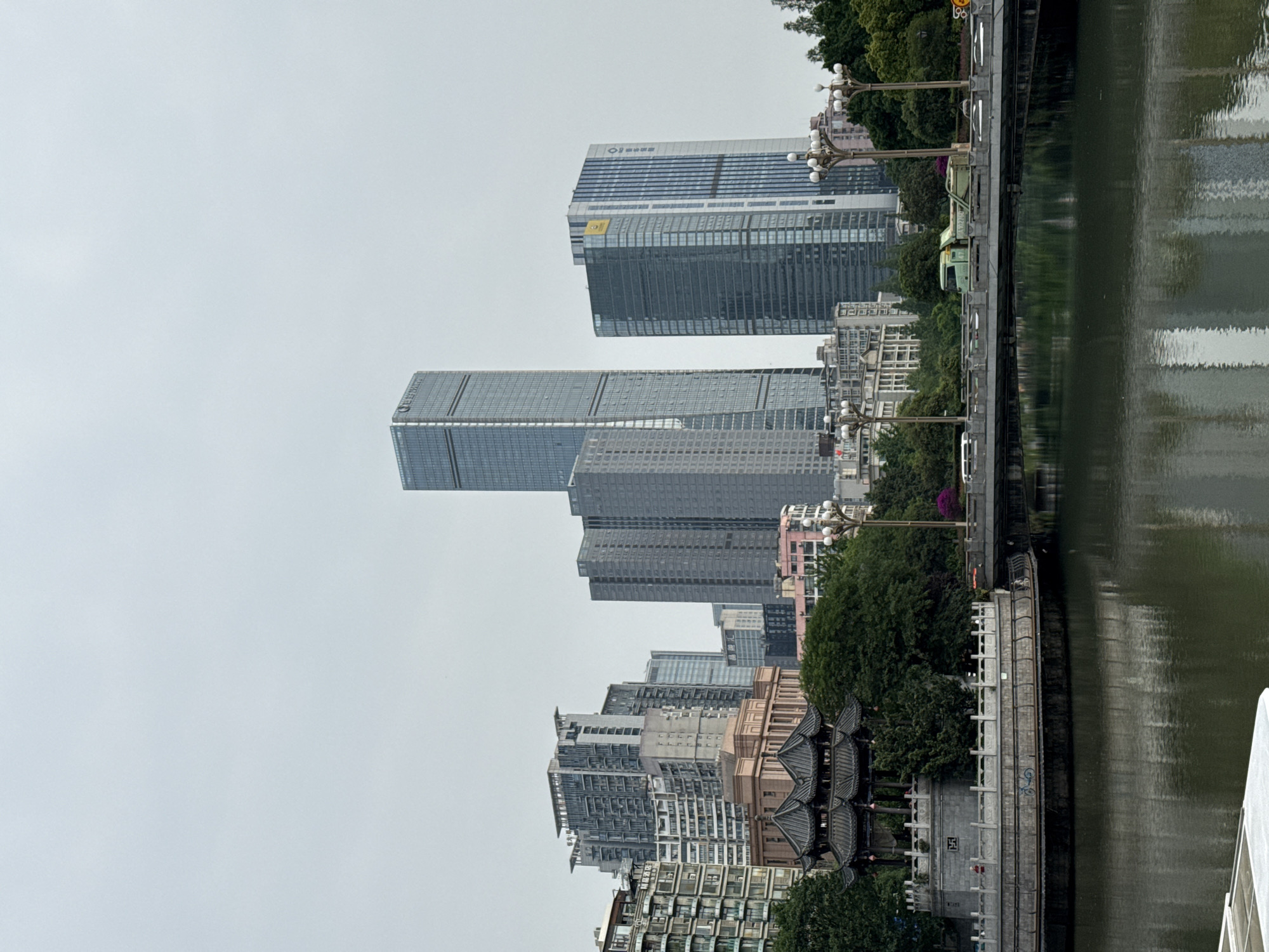 The image depicts a cityscape in Wuhou District, China. Tall, modern skyscrapers dominate the skyline. In the foreground, there is a river with reflections on the surface. A bridge spans the river, featuring vintage-style lampposts. Along the riverbank, there are trees and some traditional Chinese architectural structures. The sky is overcast, creating a muted lighting.