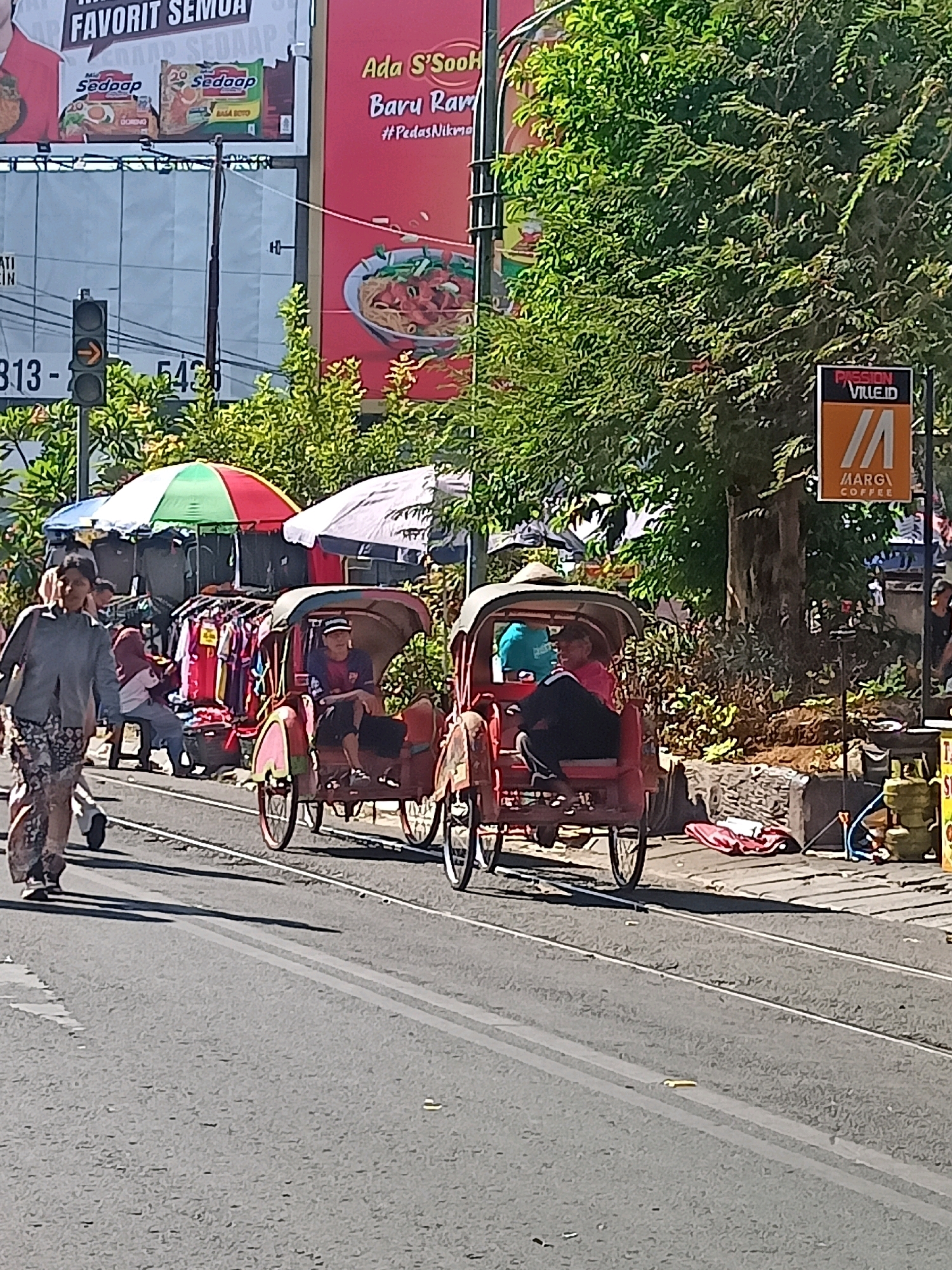 In this street scene from Surakarta, Indonesia, a woman in a patterned skirt and a gray top walks towards the left, carrying a bag. Behind her, two pedicabs are visible, each with a driver and a passenger. The pedicab on the right has a driver wearing a blue shirt and a passenger wearing a pink shirt and black pants. The pedicab on the left has a driver wearing a dark shirt and a passenger wearing a blue shirt. In the background, a street vendor is selling clothes from a stall with a red, green, and white umbrella. A traffic light with an orange arrow is visible to the left, and a billboard with Indonesian text and images of food is behind the vendor. Trees line the right side of the street, and a sign for "MARGI COFFEE" is mounted on a pole. The road appears to be paved with asphalt and has tram tracks running through it.