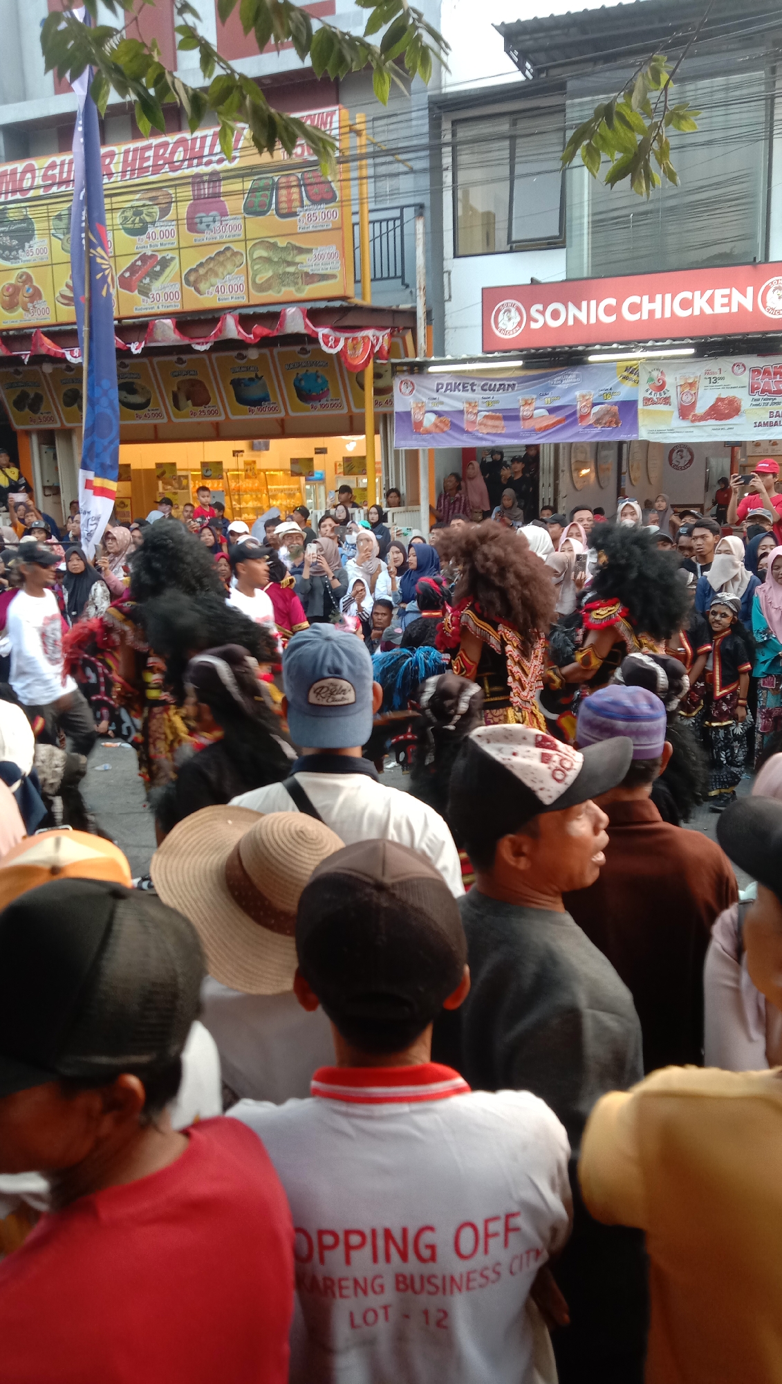This image captures a vibrant street scene in Jatisawit, Indonesia, likely during a festival or event. In the foreground, a crowd of people is gathered, with many facing away from the camera. The individuals are dressed in casual clothing, and some are wearing hats. The central focus of the image is a performance featuring two individuals in elaborate, horse-like costumes with large, dark manes and decorative attire. They appear to be the main performers, possibly engaged in a traditional dance or procession.
Behind the performers, a diverse crowd of spectators is visible, many of whom are holding up their phones to capture the event. They are arranged in front of various storefronts and banners. One prominent banner displays the logo "SONIC CHICKEN" with Indonesian text, indicating a commercial establishment. Other banners advertise food items, with prices listed in Rupiah, suggesting a food or street market setting. The buildings in the background appear to be typical of an urban environment, with shop fronts and overhead wires. The overall atmosphere seems lively and festive, with a strong sense of community engagement. The lighting suggests it is daytime, possibly late afternoon.