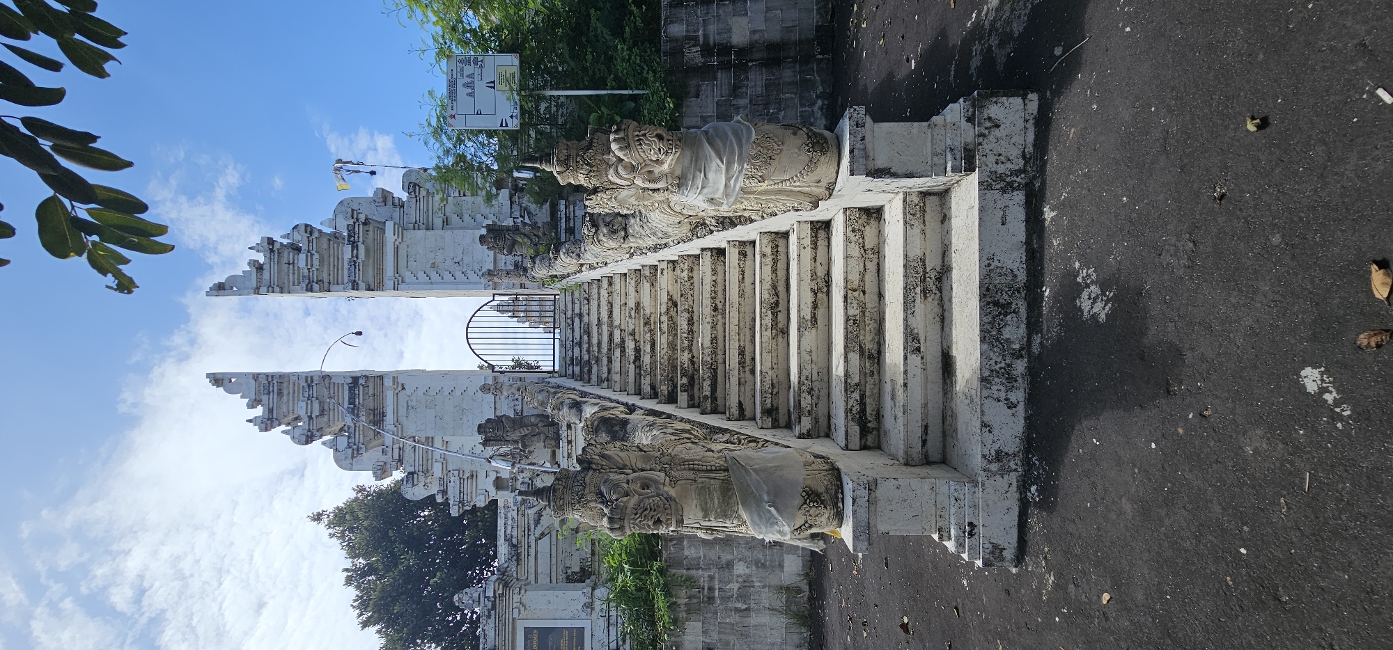 The image shows a traditional Balinese gate and staircase in Kuta Selatan, Indonesia. The gate, constructed of white stone, features intricate carvings and leads to a set of stairs flanked by stone statues. A metal gate is visible at the top of the stairs. The surrounding area includes trees, greenery, and an asphalt surface. The scene is bathed in daylight, with a clear blue sky and scattered clouds overhead.
