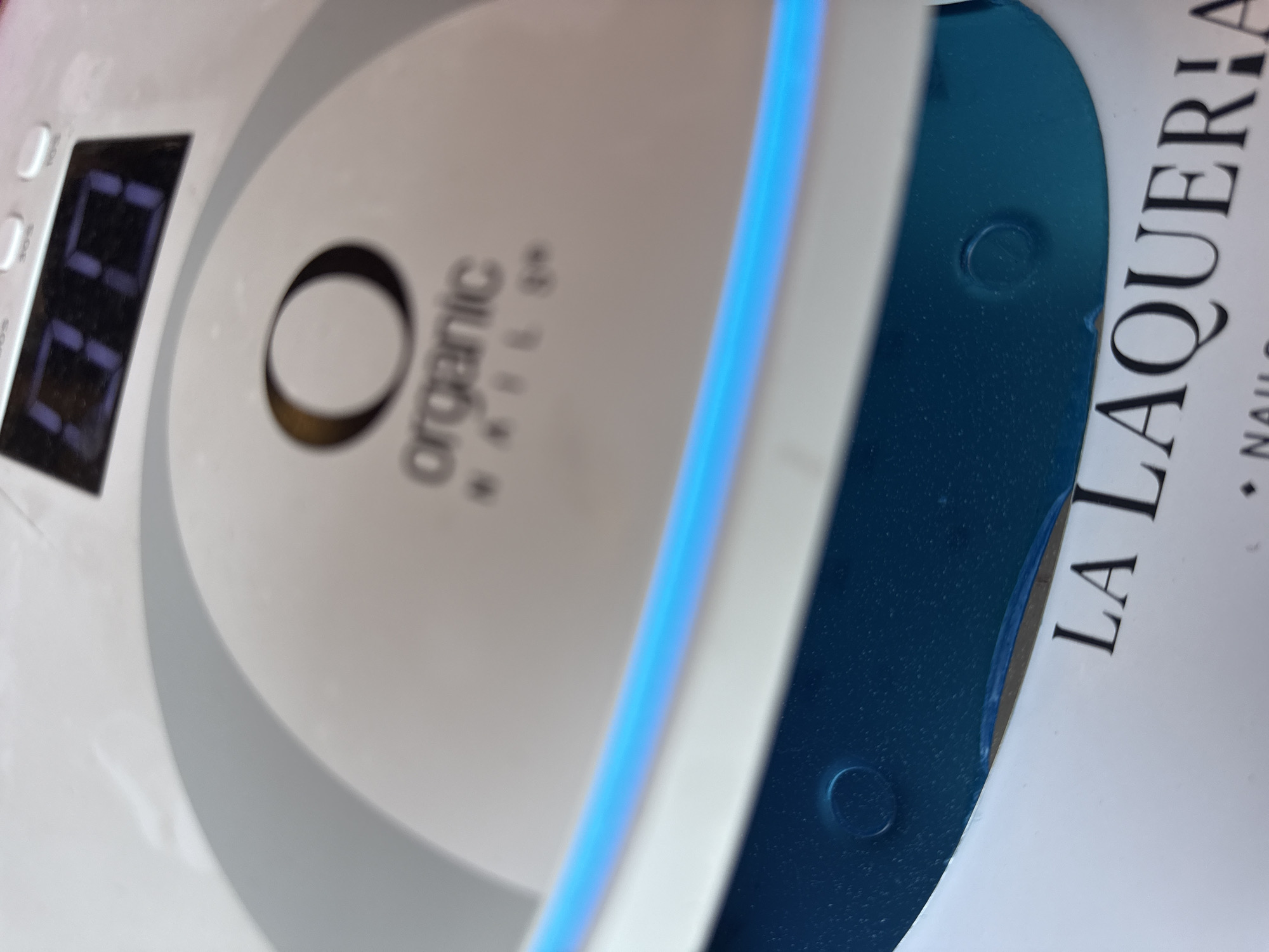 The image presents a close-up, angled view of a white UV/LED nail lamp, likely in use within a nail salon environment in Puebla, Mexico. The lamp features a digital display that reads "00", suggesting it is either in standby mode, awaiting a timer setting, or has just completed a cycle. A bright blue light, indicative of UV or LED curing technology, is visible emanating from a slot on the device. To the left of the display, faint text "30S" and "60S" can be discerned near buttons, likely indicating preset timing options for nail polish curing.
The brand logo "O organic NAILS" is prominently displayed on the white surface of the lamp. Adjacent to the lamp, a deep blue object, possibly a tray, mat, or another piece of nail salon equipment, is partially visible. On this blue surface, the text "LA LAQUERIA" is clearly printed in large black capital letters, followed by "NAILS", further identifying the setting as a professional nail salon. The composition focuses on the equipment and branding, suggesting an ongoing or imminent nail treatment activity.